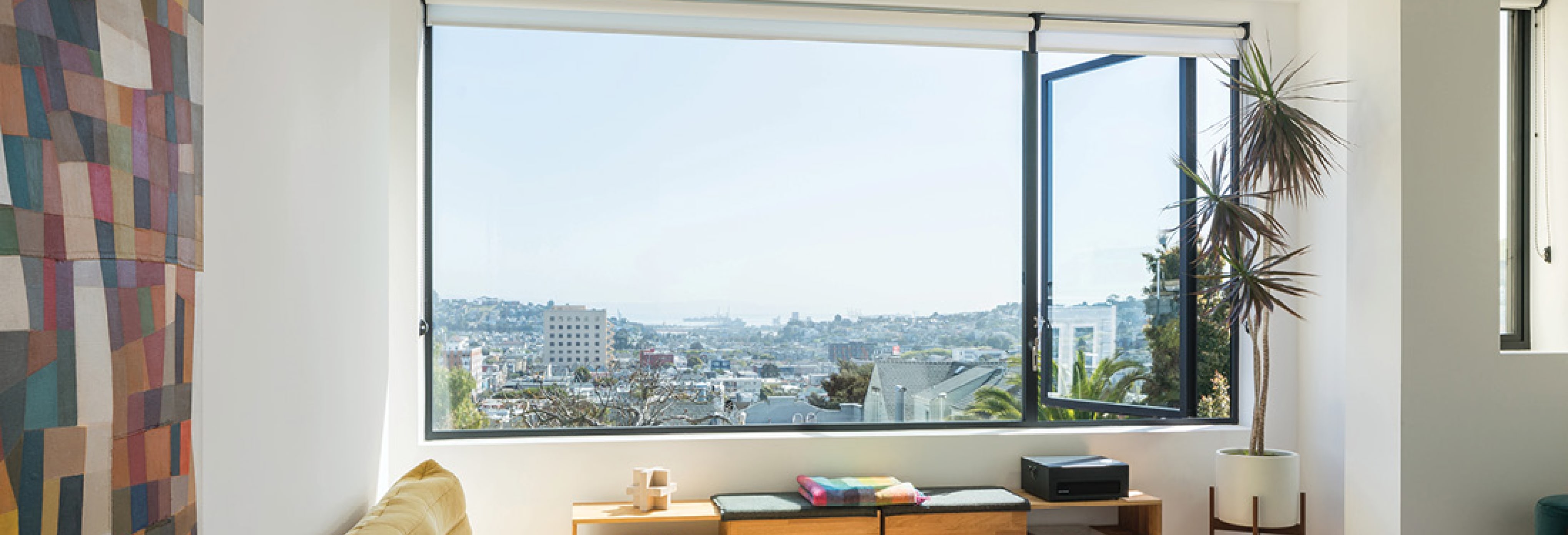 Modern bedroom with large picture window offering panoramic city views, mustard yellow tufted lounge chair, colorful geometric wall art, wooden storage bench with cushions, and potted indoor plant.
