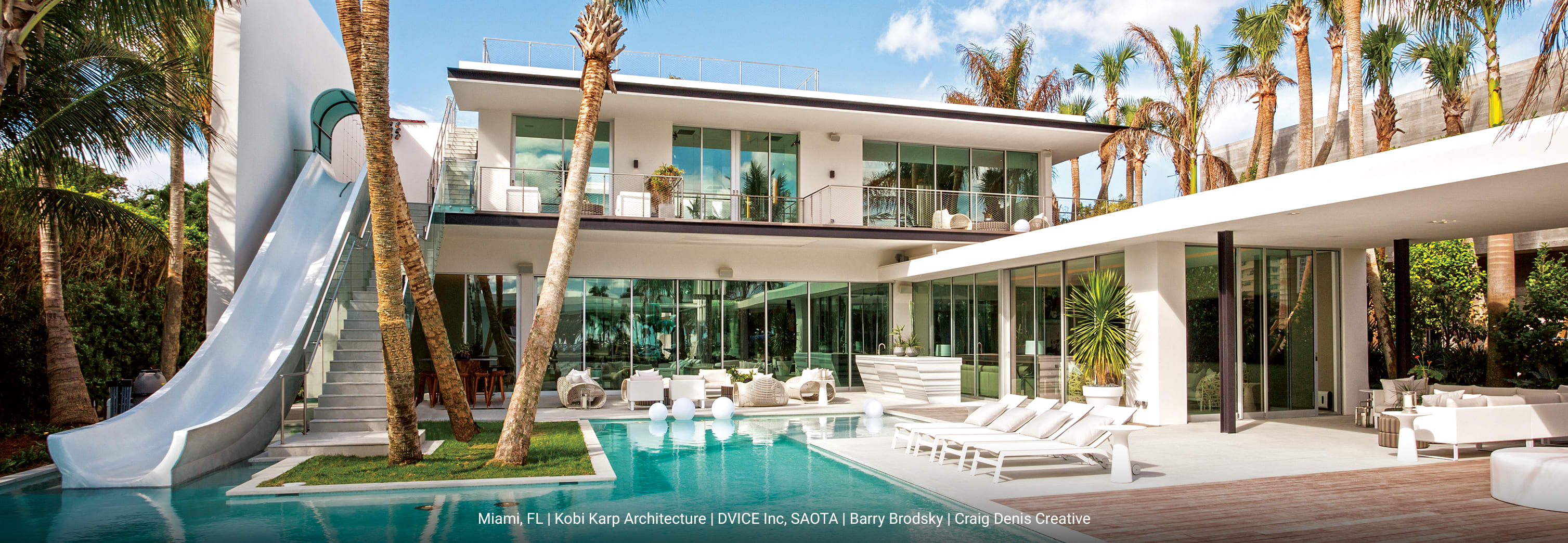 Luxury modern waterfront home with minimalist concrete exterior, expansive floor-to-ceiling glass walls, wraparound balcony with glass railing, and outdoor living space featuring lounge seating and swimming pool surrounded by palm trees.