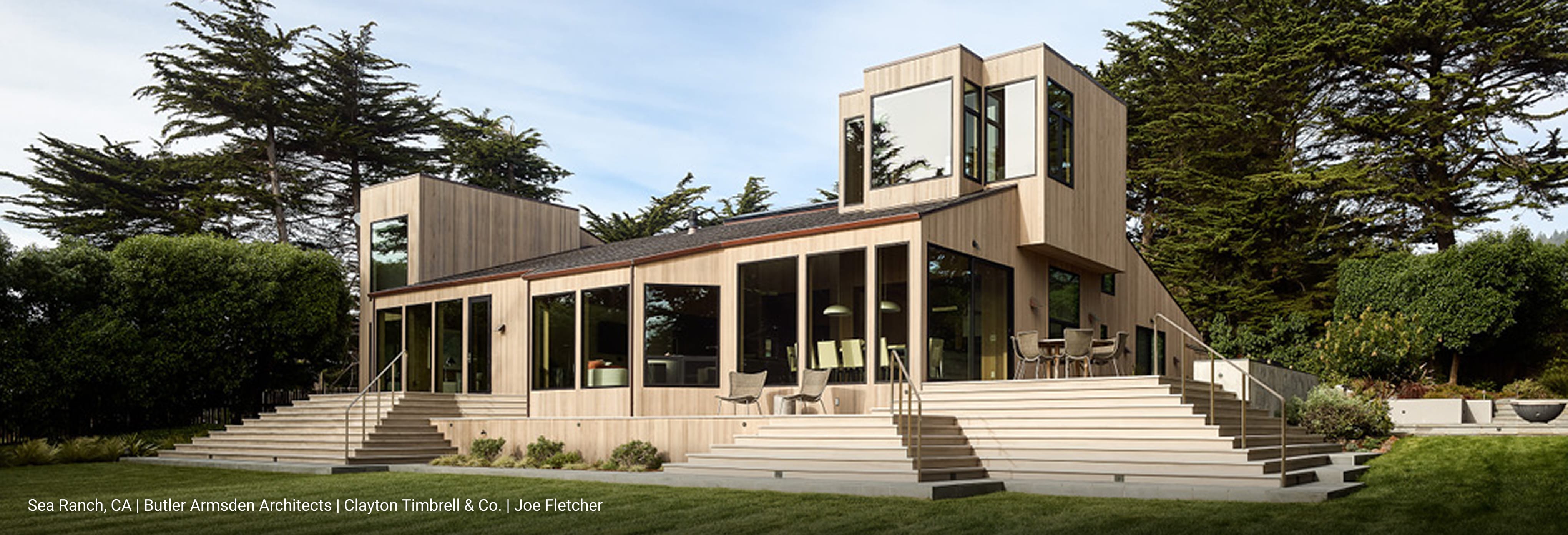 Contemporary two-story home with natural wood siding, large glass windows, and sloped roof design, featuring expansive outdoor steps leading to a landscaped lawn surrounded by tall evergreen trees.