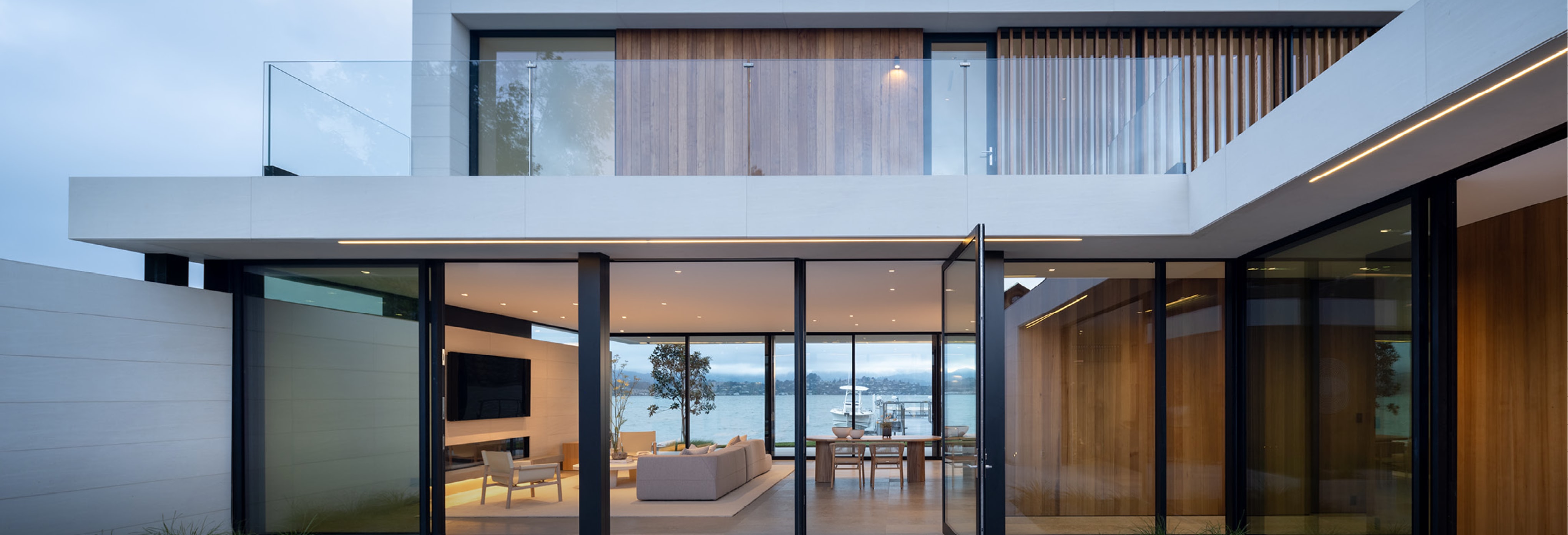 Contemporary two-story home with minimalist design, white exterior walls, glass balcony railings, vertical wooden slats, and floor-to-ceiling glass doors opening to an outdoor terrace overlooking water views.
