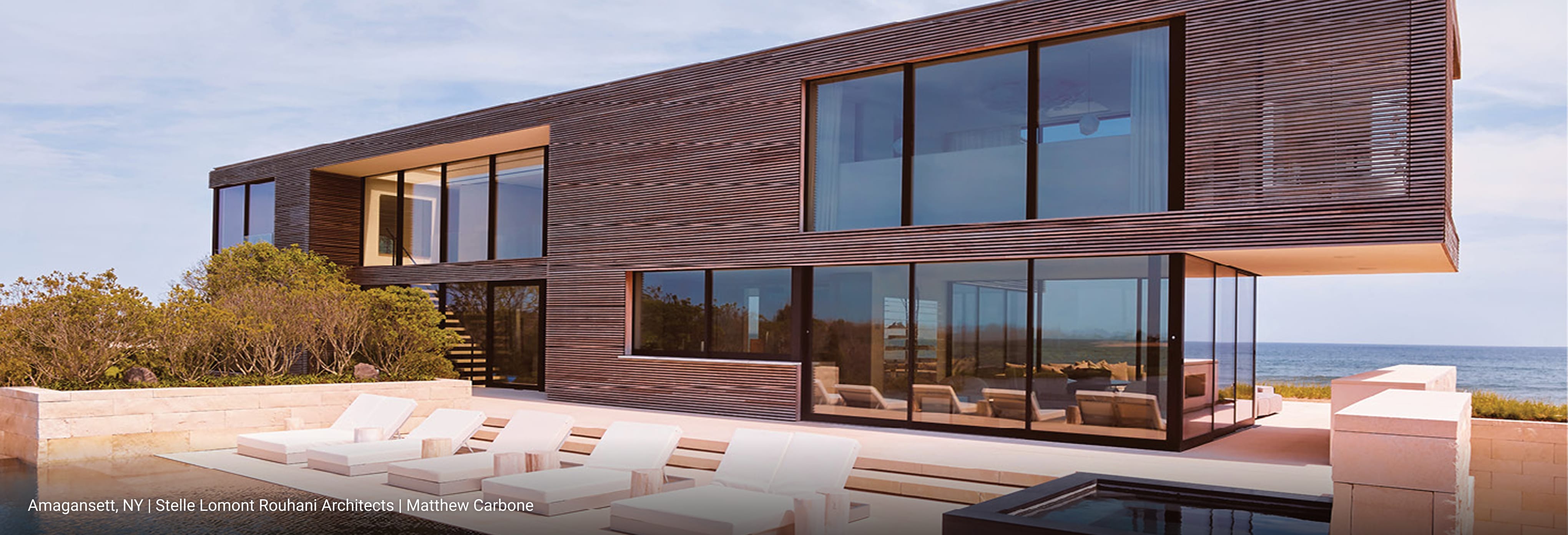 Contemporary oceanfront home with horizontal wood slat siding, expansive floor-to-ceiling glass windows, cantilevered upper level, and outdoor lounge area with white sunbeds overlooking the Atlantic.