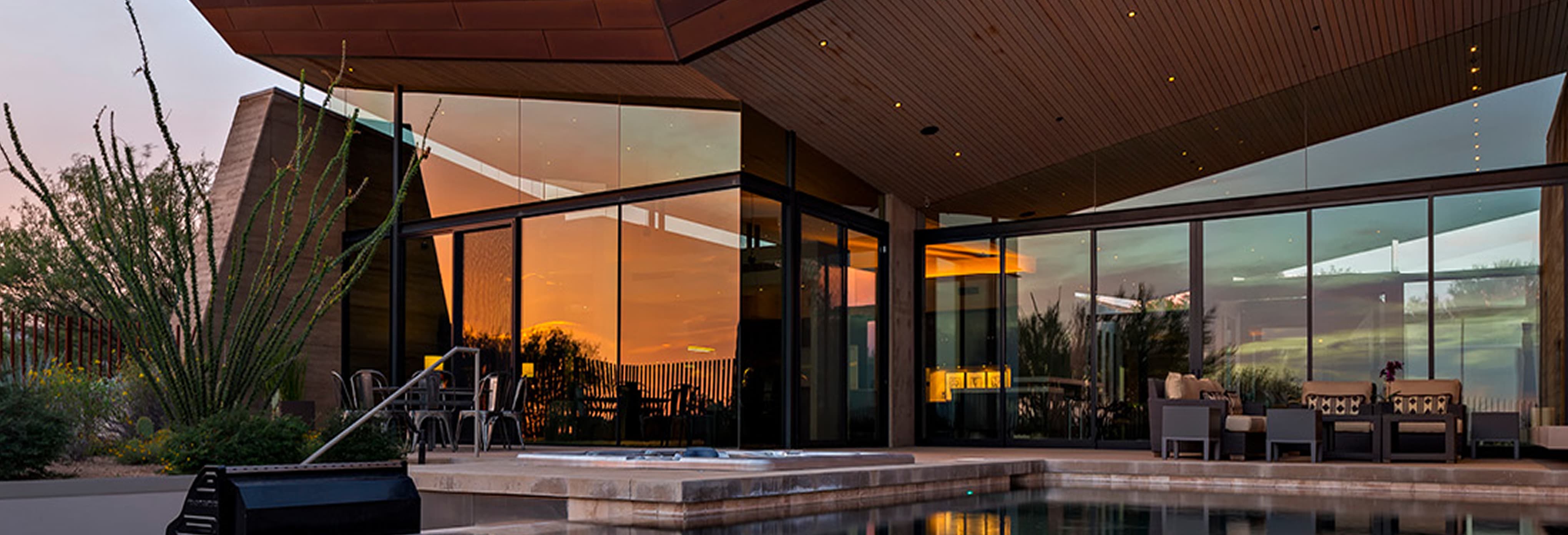 Contemporary desert home with floor-to-ceiling glass walls, sleek wooden ceiling, outdoor seating area, and pool reflecting a vibrant sunset.