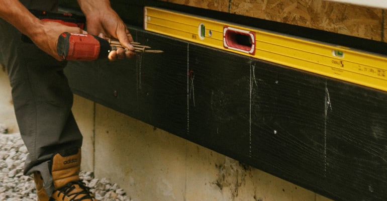 Worker using a drill and checking alignment with a yellow level on a black exterior board during construction.