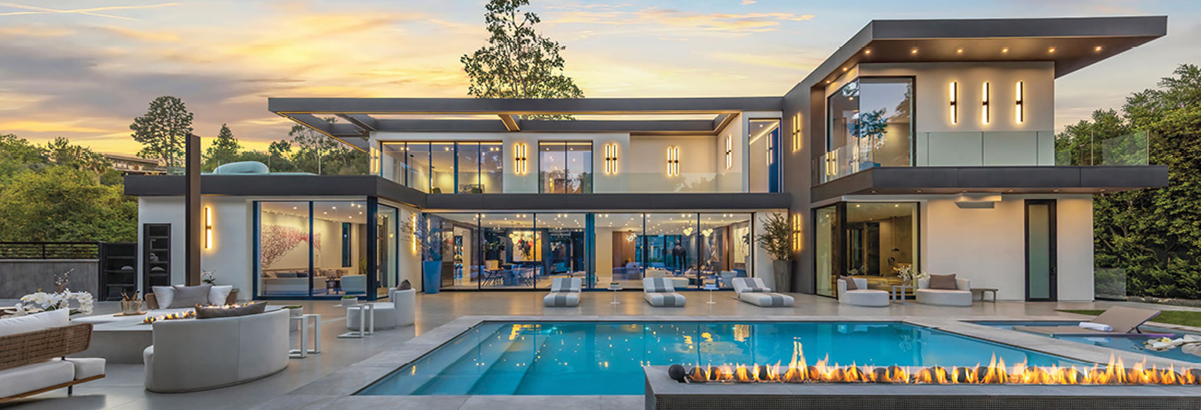 Luxury modern two-story home with expansive glass walls, outdoor lounge seating, infinity pool, and fire feature at sunset.