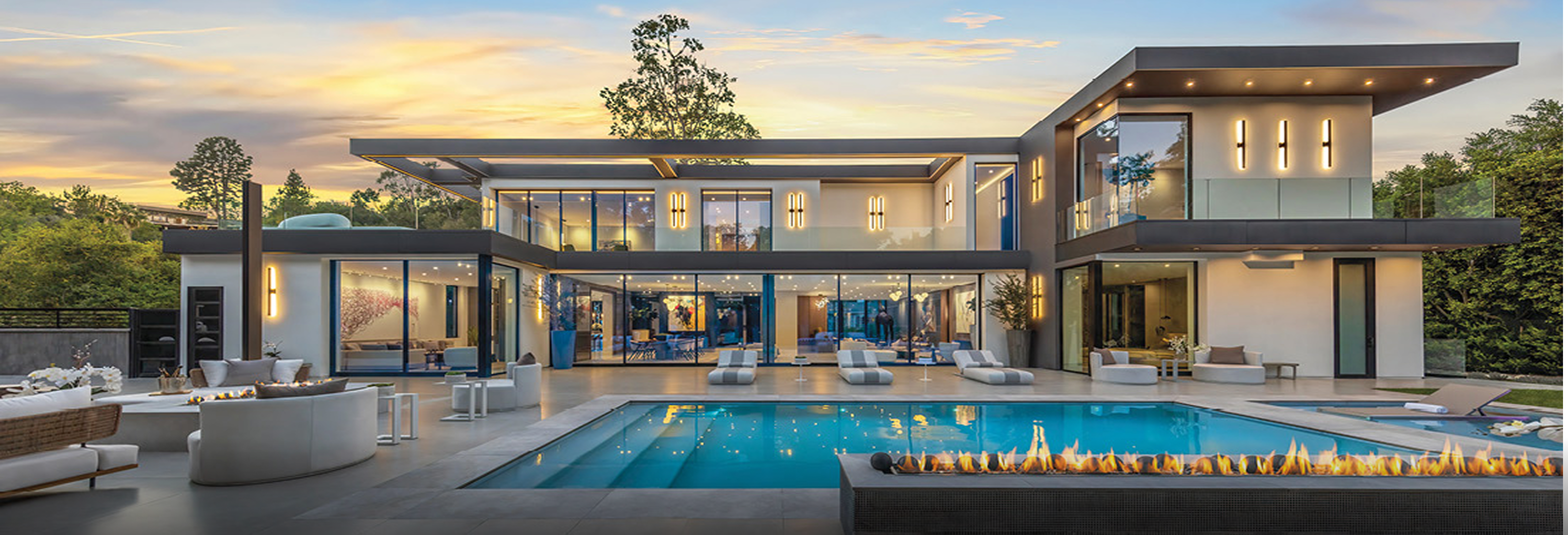 Luxury modern two-story home with floor-to-ceiling glass walls, sleek flat roof design, outdoor lounge seating, and large swimming pool with fire feature at sunset.