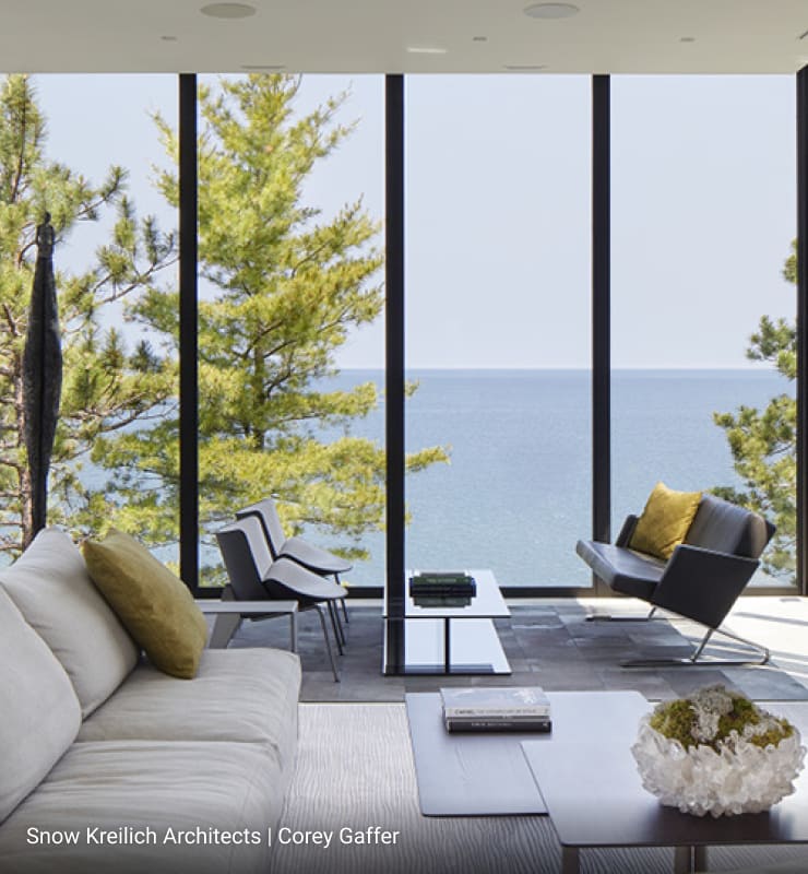 Modern living room with floor-to-ceiling glass walls offering panoramic lake views, minimalist furniture including light beige sofa and black lounge chair, sleek coffee tables, and natural stone accent wall.