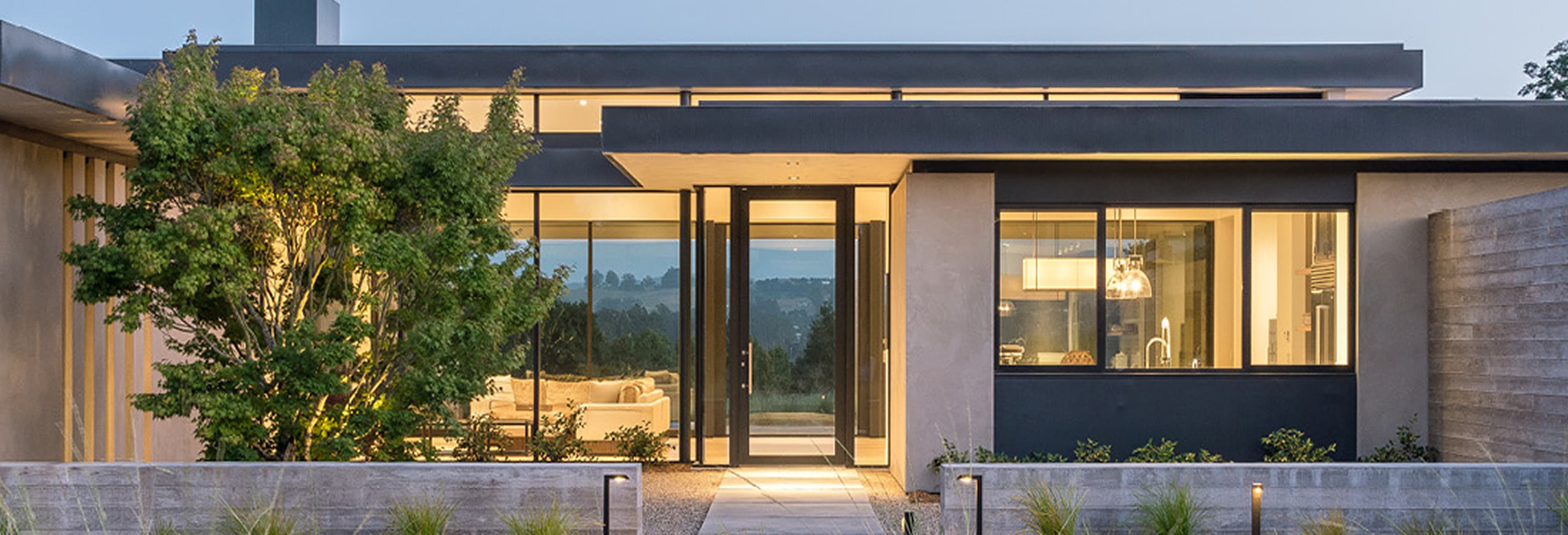Modern single-story home with clean architectural lines, large glass windows, minimalist landscaping, and warm interior lighting at dusk.