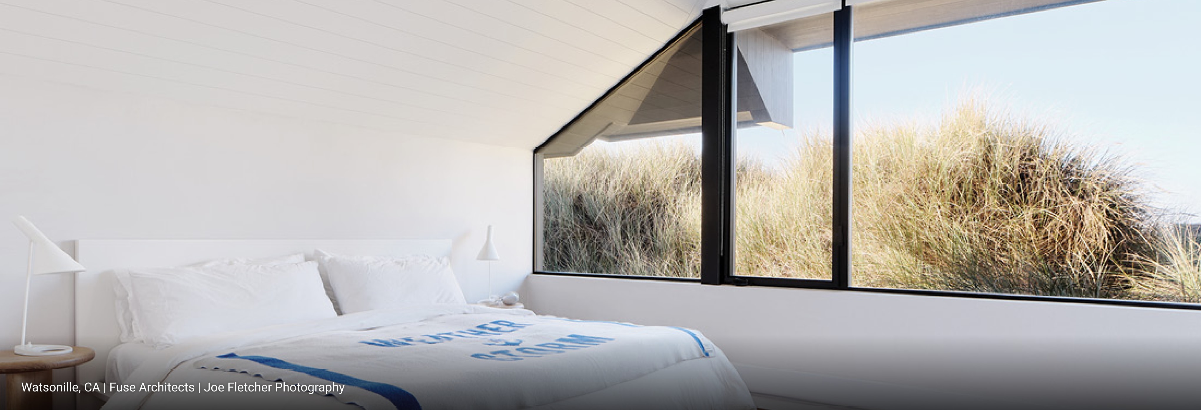 Minimalist coastal bedroom with white bed frame, crisp white bedding, light wood side table, modern white lamp, and large black-framed windows overlooking sand dunes and natural grasses.