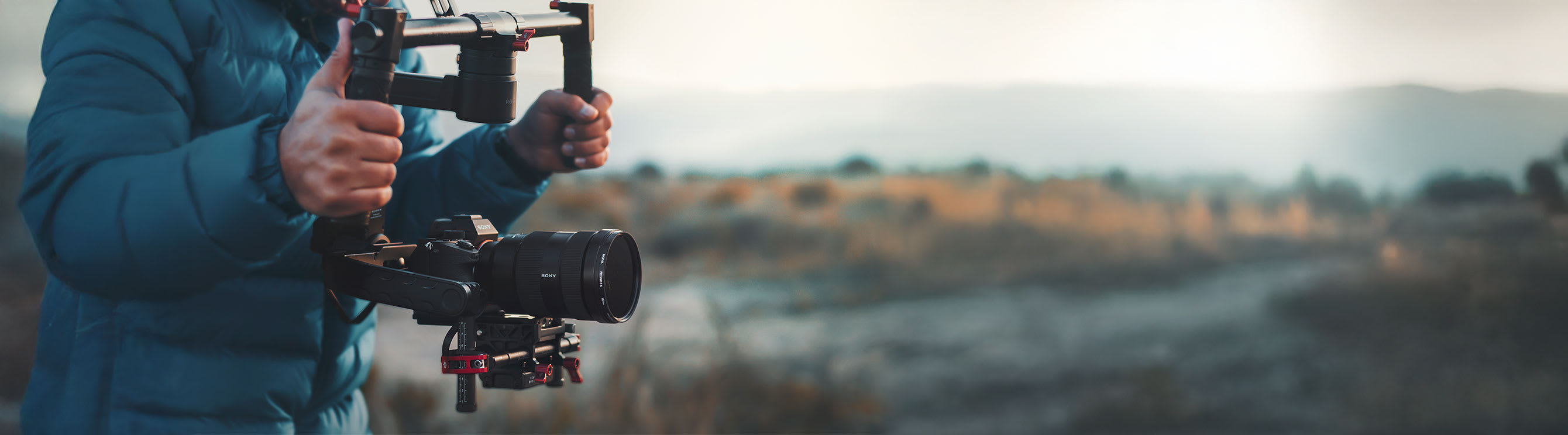 Person standing in field, aiming a high-end camera, wearing a blue puffer jacket