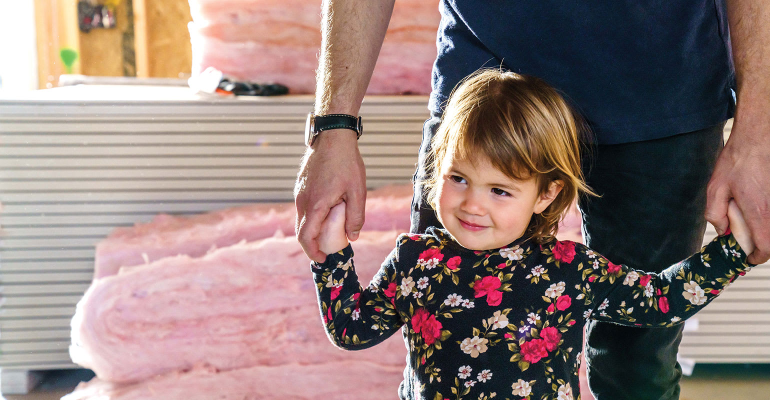 Photo of an adult holding the hands of a toddler with a house and insulation in the background