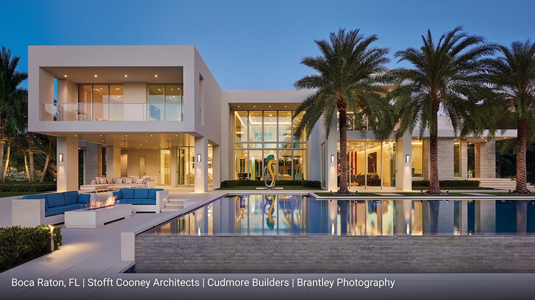 Luxury modern home in Boca Raton, Florida featuring flat roof design, white stucco exterior, expansive floor-to-ceiling glass windows, and outdoor living space with fire pit lounge, palm trees, and infinity-edge pool.