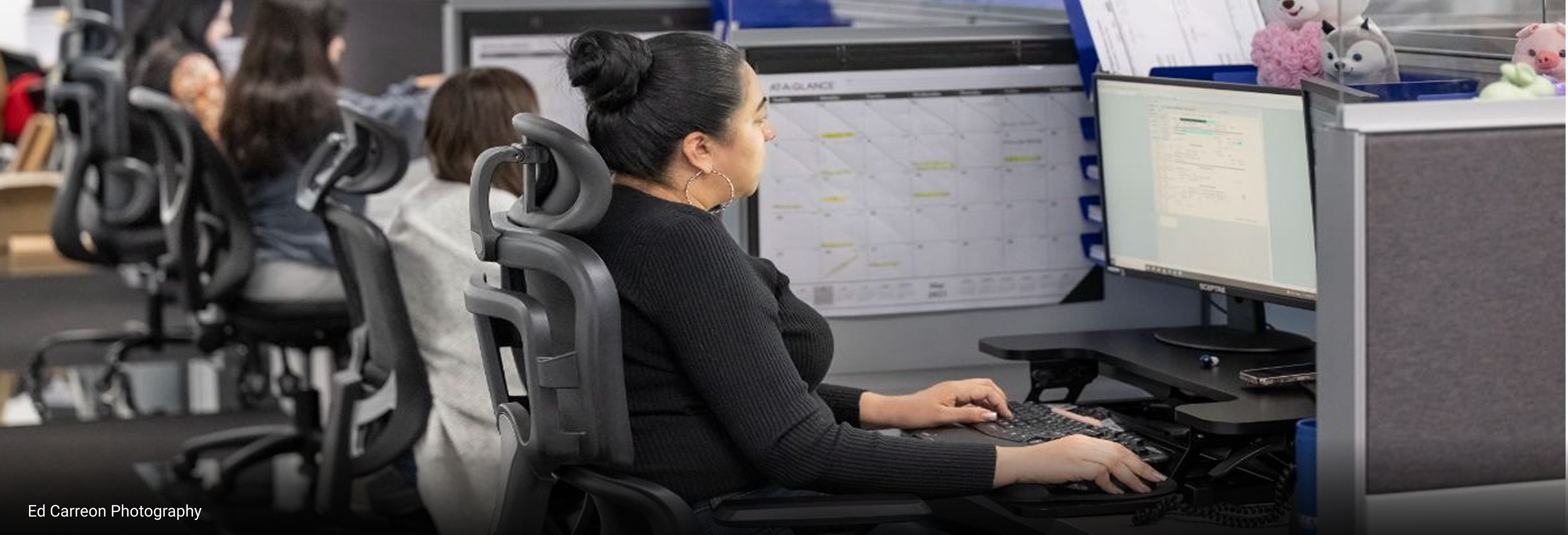 Office workspace with ergonomic chairs and computer monitors displaying scheduling and project management tools, featuring organized cubicles with calendars and personal desk accessories.