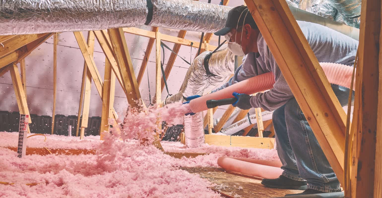 A man dressed in proper safety gear such as, eyewear protection, gloves, face mask, and long-sleeved, loose-fitting clothing, uses a hose to install PROPINK® L77 PINK® Fiberglas™ Unbonded Loosefill Insulation as part of the process for the PROPINK Complete™ Insulation System.