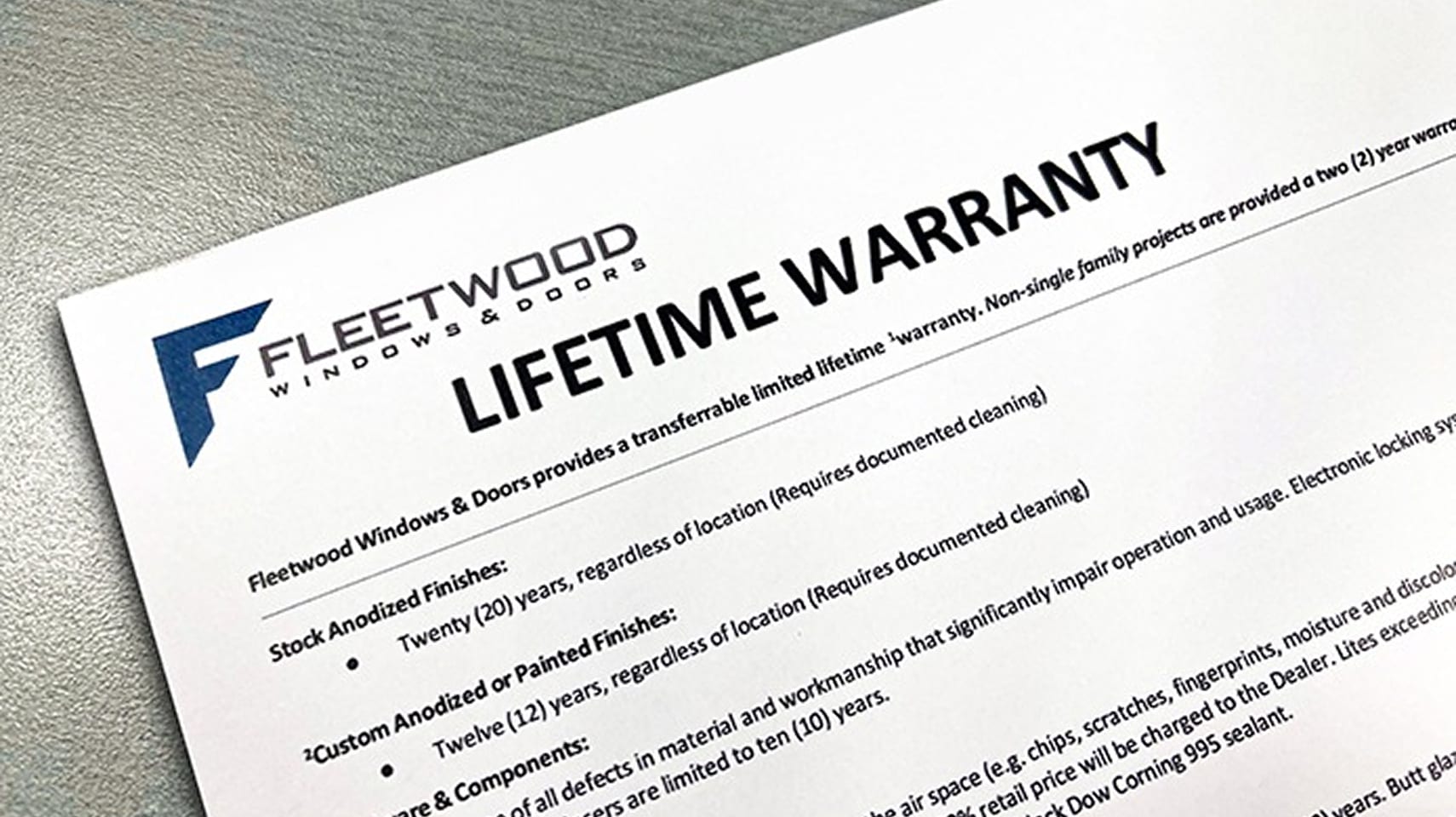 Fleetwood lifetime warranty document detailing coverage for anodized finishes, painted finishes, hardware components, and insulated glass, displayed on a gray textured surface.