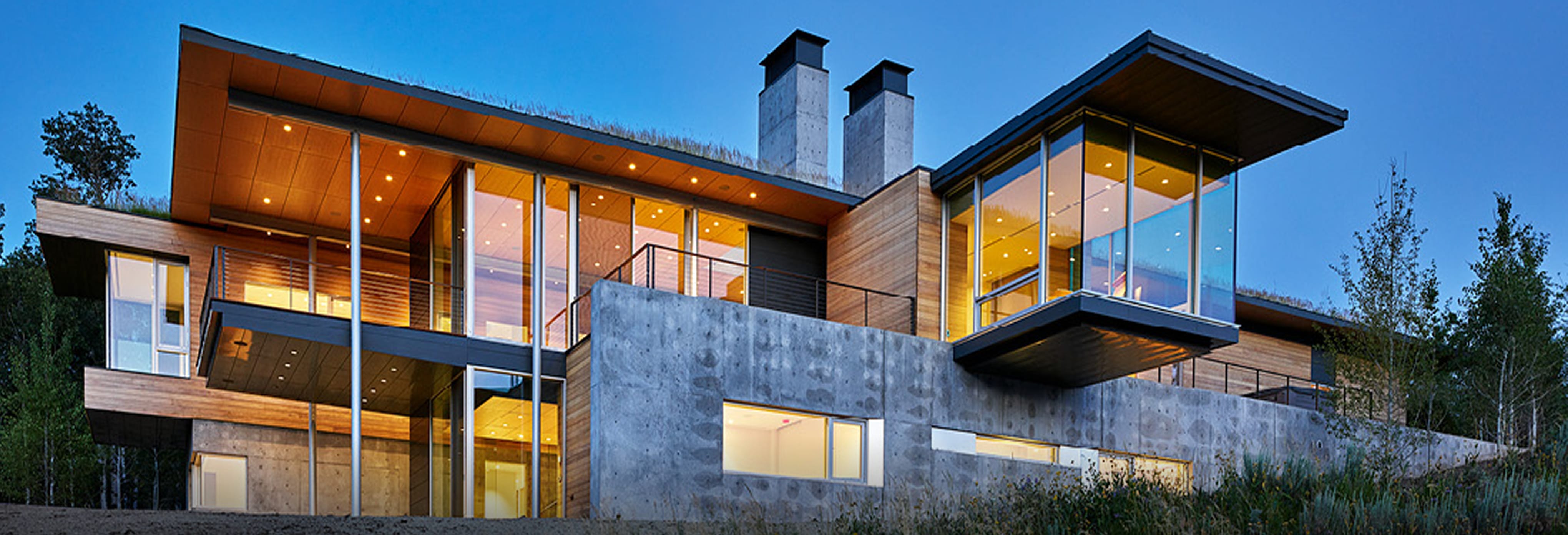 Contemporary hillside home with cantilevered design, flat rooflines, exposed concrete walls, natural wood siding, and expansive floor-to-ceiling glass windows illuminated at dusk.
