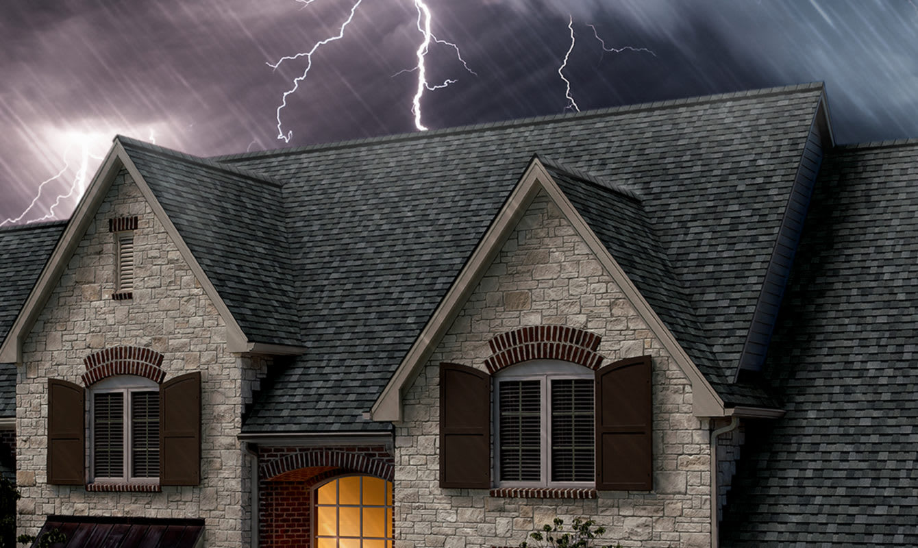 Photo of the top of a roof with gray shingles set against a dark, stormy sky with lightning