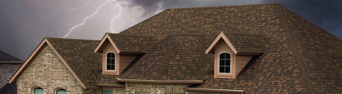 Tan colored house with brown shingles set against a dark, stormy sky with lightning flashing