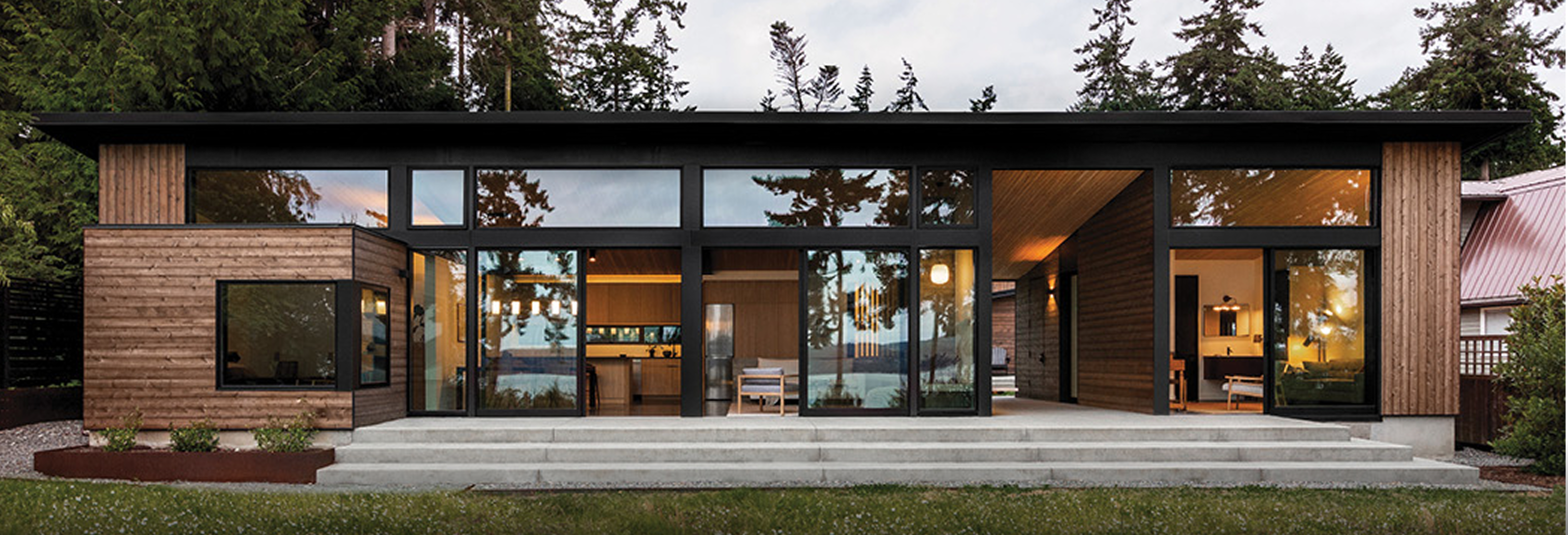 Contemporary single-story home with wood siding, large glass windows, flat roof, and open interior layout surrounded by tall evergreen trees.