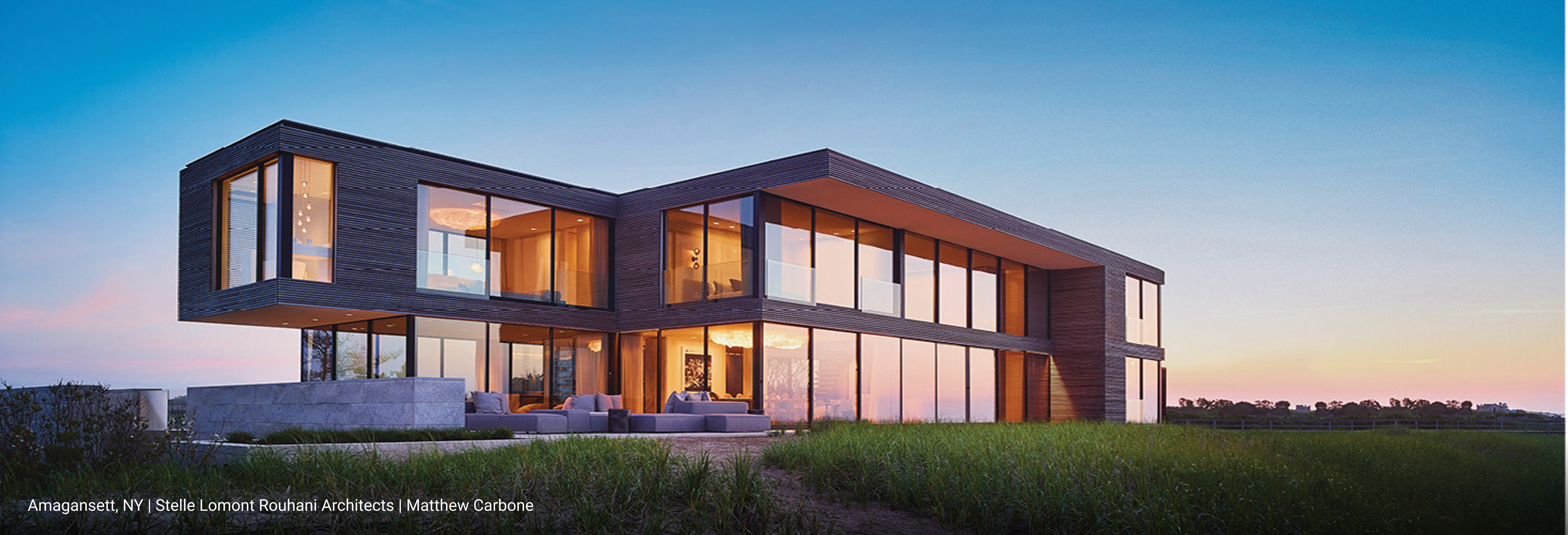 Luxury modern home in Amagansett, New York featuring cantilevered design, dark horizontal wood siding, expansive floor-to-ceiling glass windows, and open-concept layout overlooking coastal landscape at sunset.