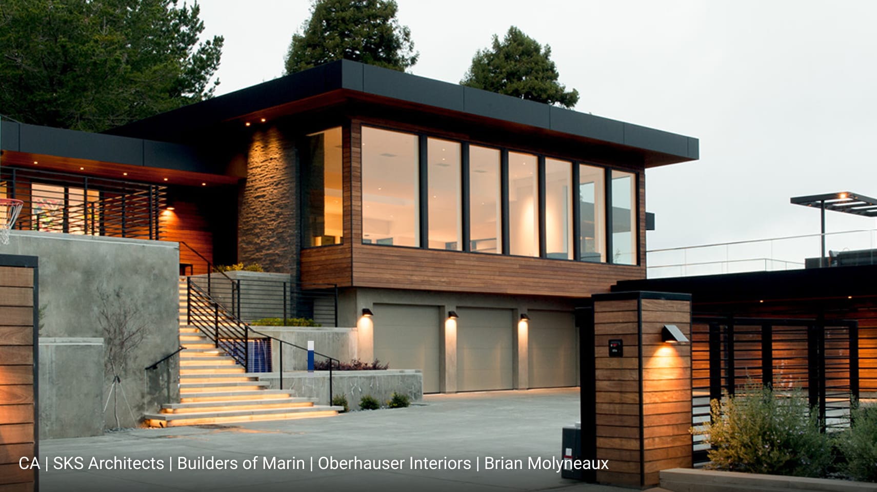 Modern California home with flat roof design, natural wood siding, stone accent walls, and expansive floor-to-ceiling glass windows, featuring illuminated entry steps and three-car garage.
