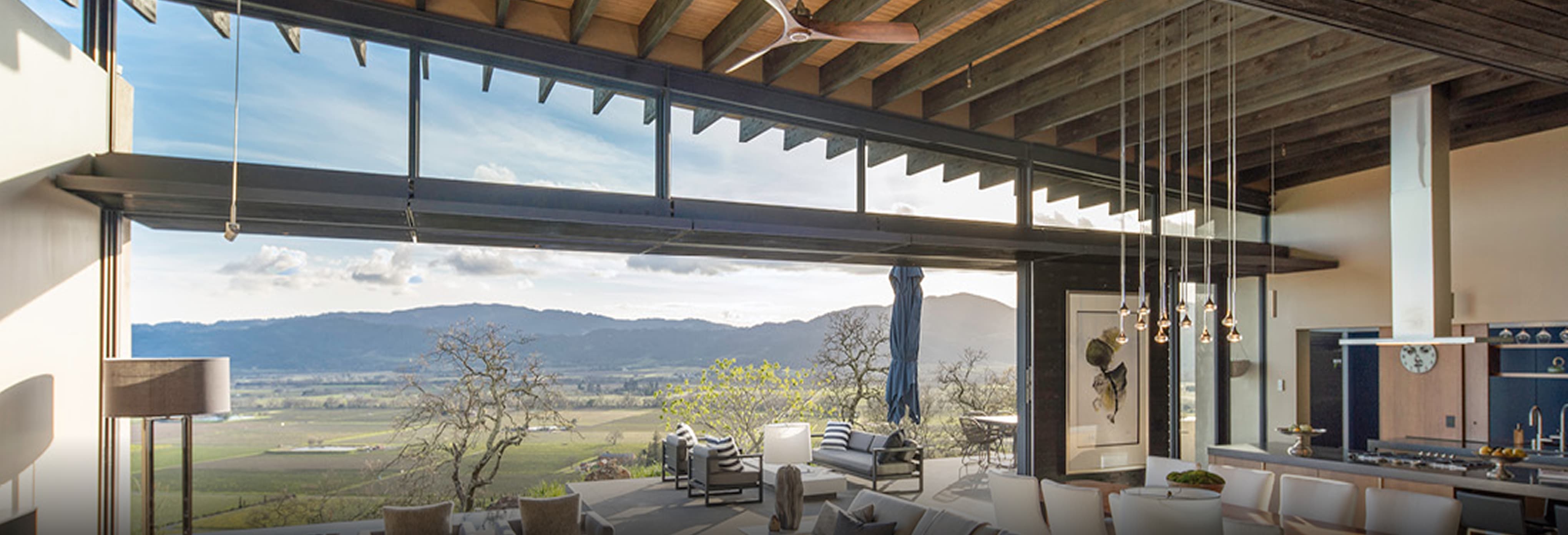 Luxury open-concept living and dining area with exposed wood ceiling beams, floor-to-ceiling glass walls, and folding glass doors opening to a terrace with panoramic vineyard views, featuring modern furniture and pendant lighting.