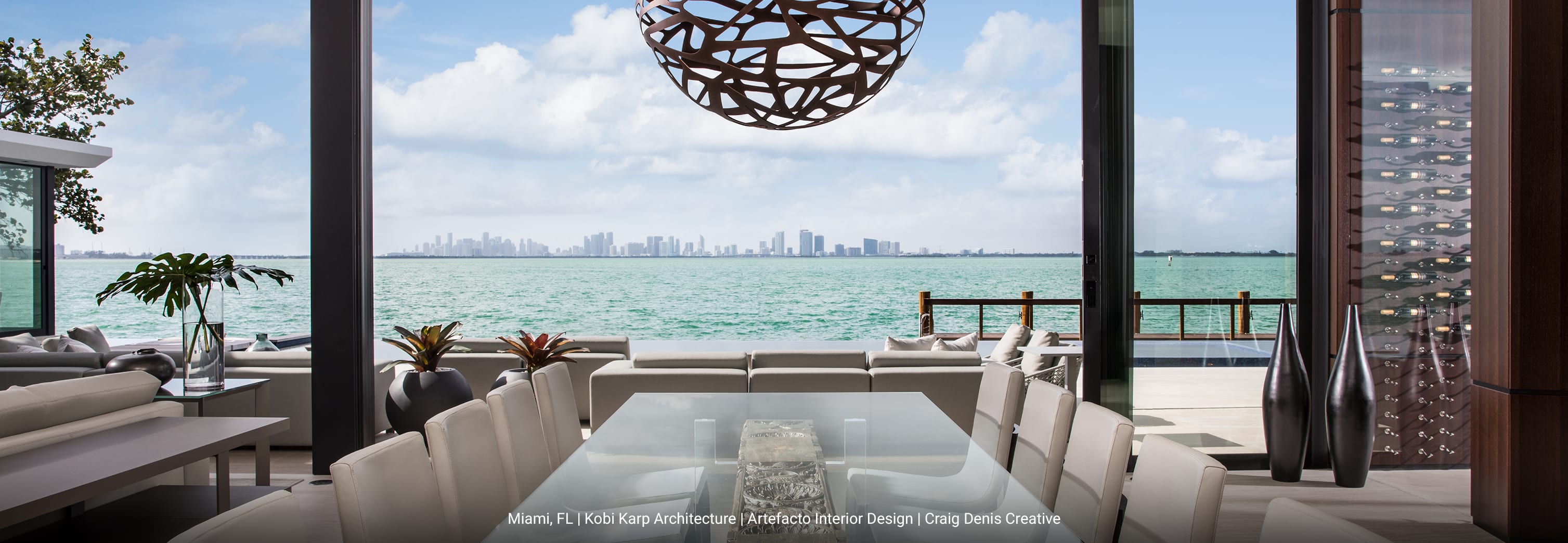 View from a dining room in a luxury waterfront modern home, featuring a glass table with white chairs, overlooking turquoise ocean water and the Miami skyline under a blue sky with white clouds.