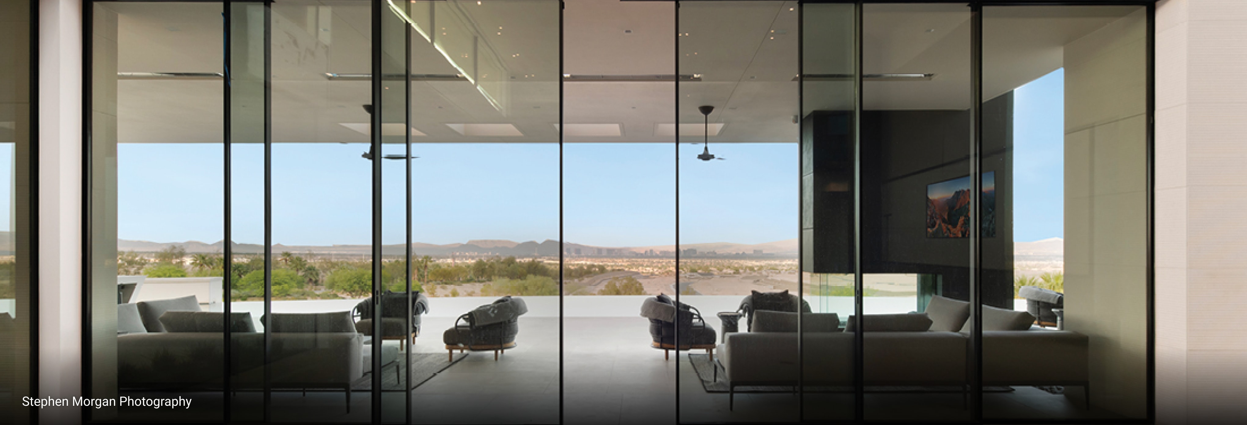 Modern living room with sleek glass walls, minimalist furniture, and panoramic desert landscape views through floor-to-ceiling windows.