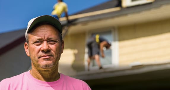 Roofing contractor wearing a pink shirt and a ball cap standing in front of a yellow house with roofers on the roof.