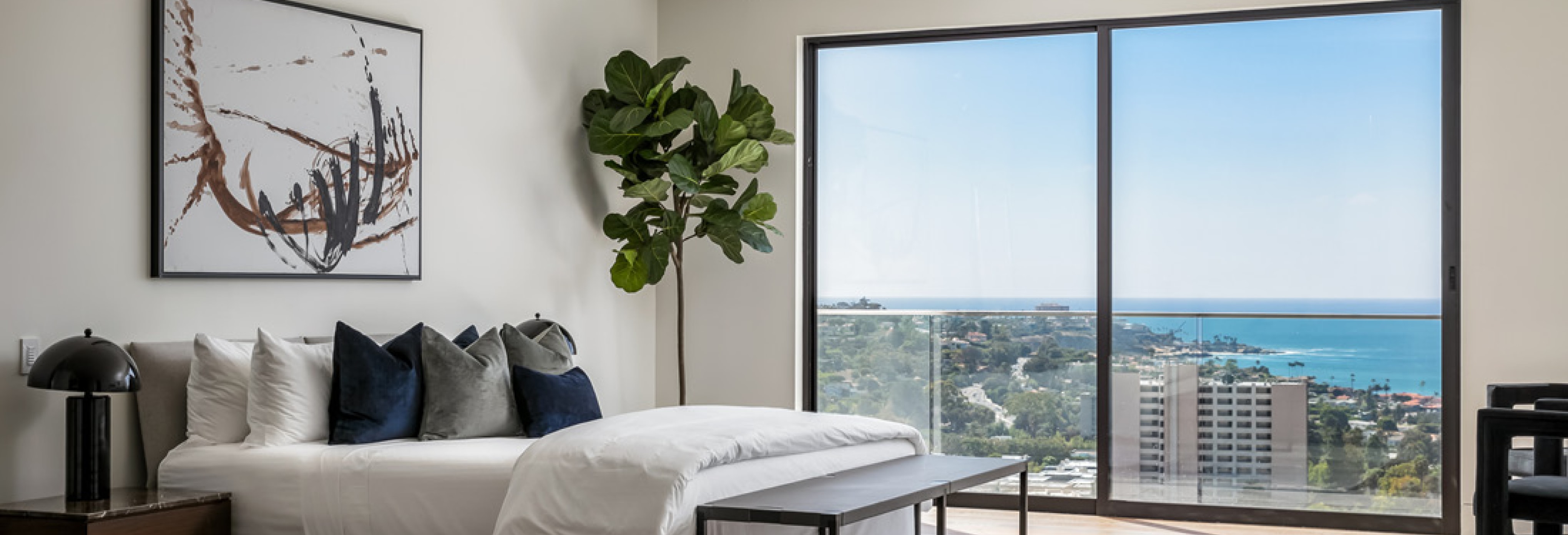 Modern bedroom with minimalist decor, white bedding, abstract wall art, potted plant, and sliding glass doors showcasing panoramic ocean views.