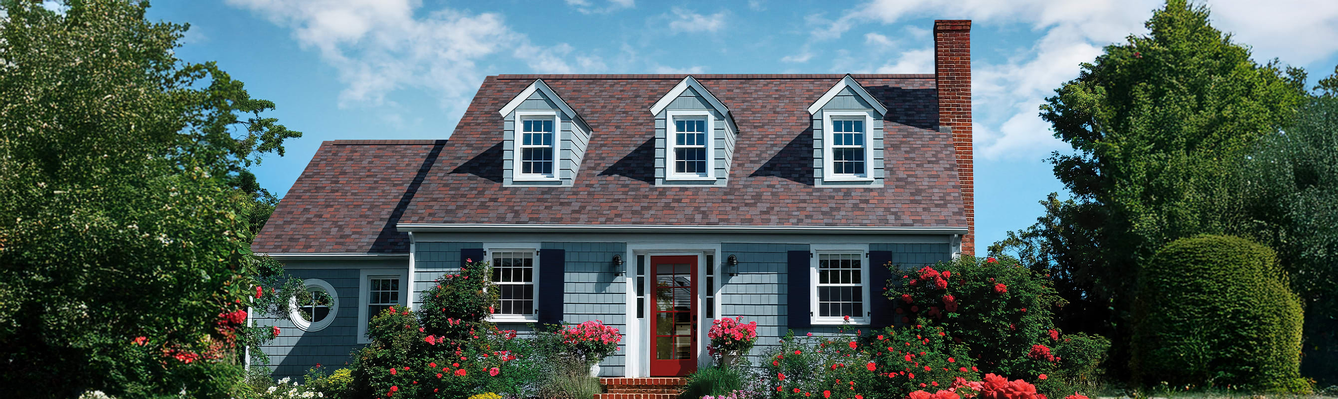 Blue grey house with Owens Corning Duration Designer shingles in Merlot, surrounded by a blue sky and shrubbery with spring flowers.