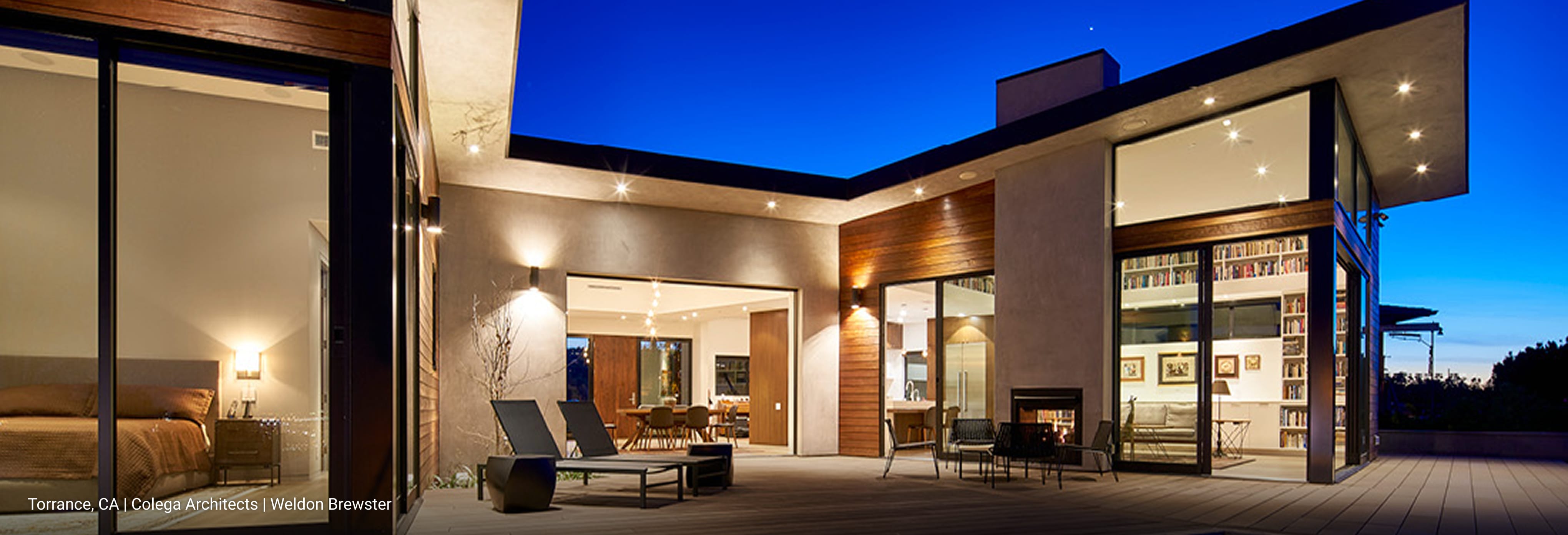 Luxury modern home in Torrance, California featuring flat roof design, natural wood siding, stucco walls, and expansive floor-to-ceiling glass windows, with illuminated outdoor lounge and open-concept interior visible at dusk.