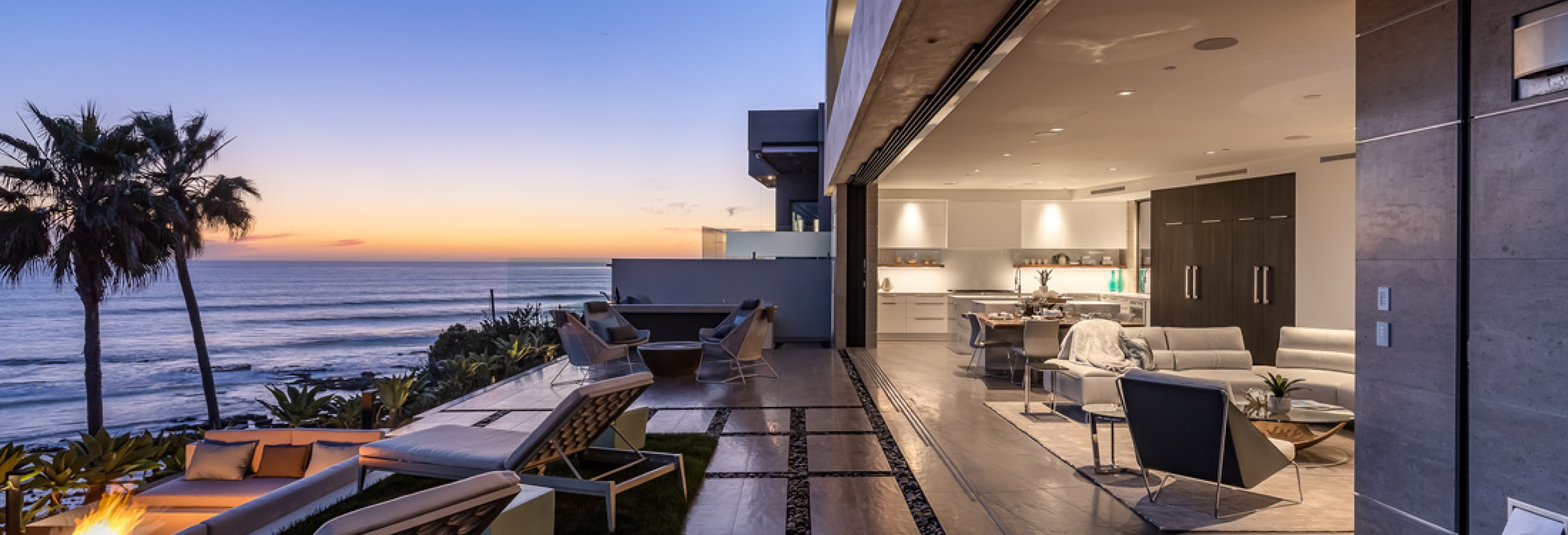 Luxury oceanfront home with open-concept living area, modern kitchen, and outdoor terrace featuring lounge chairs and fire pit overlooking sunset views of the Pacific.