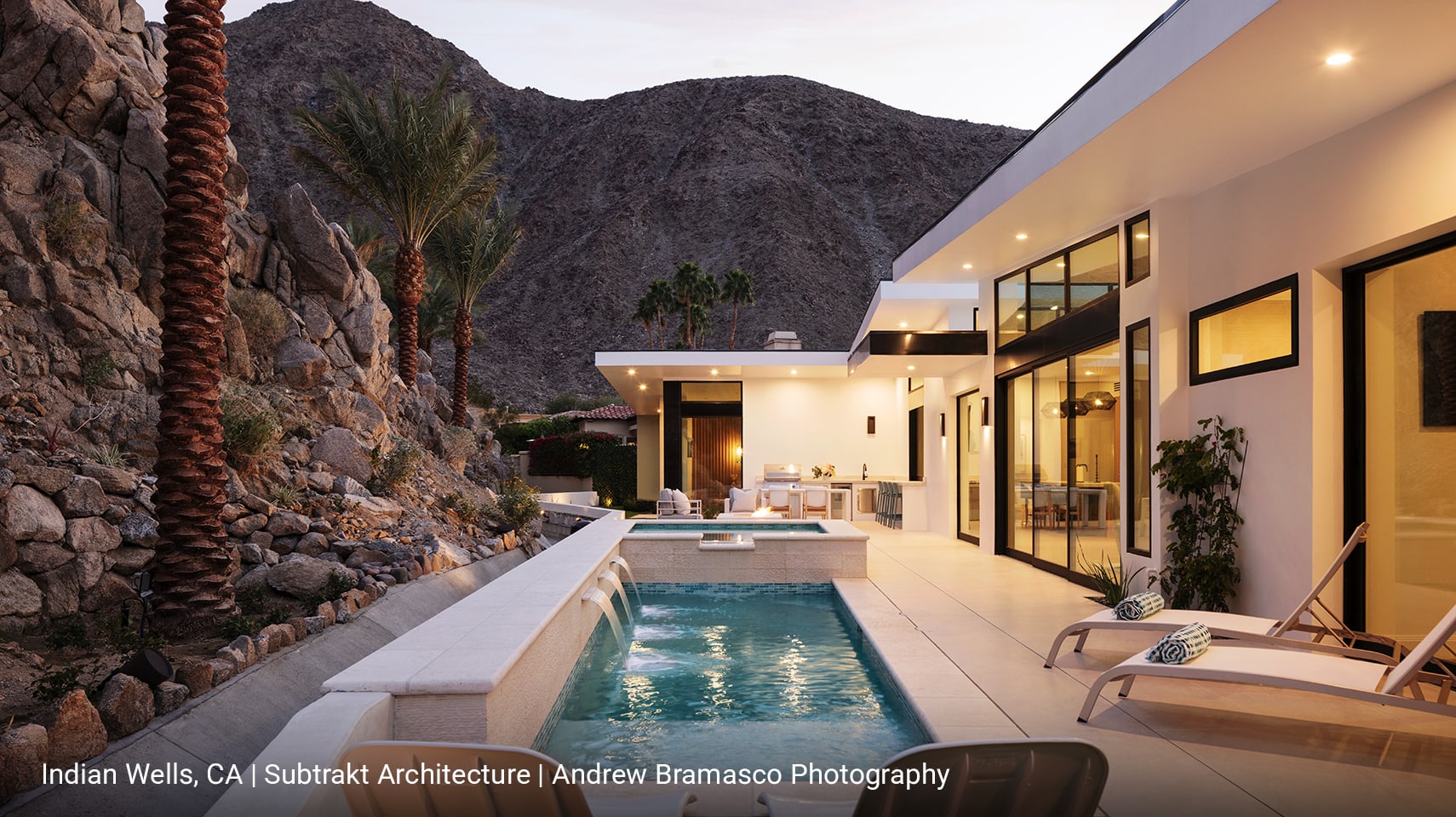 Luxury desert backyard with expansive black-anodized sliding glass doors opening to a modern pool featuring cascading waterfalls, an integrated hot tub, and sleek lounge chairs framed by arid landscape.