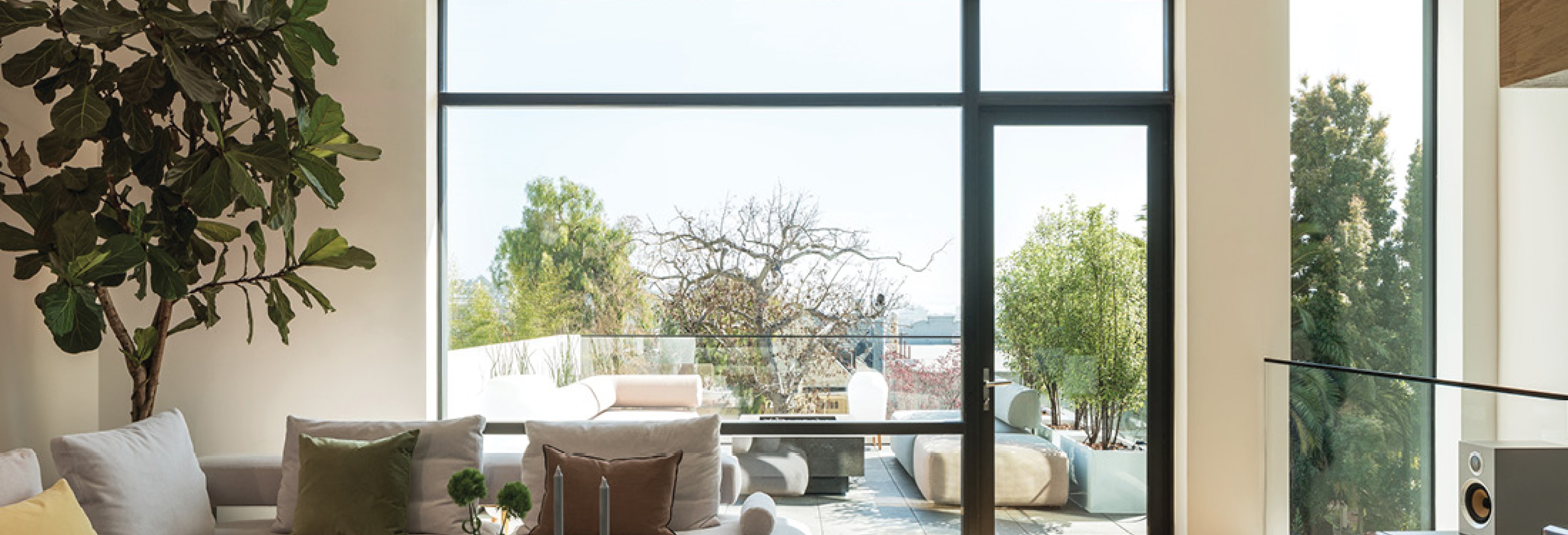 Modern living room with large floor-to-ceiling glass windows and door opening to a balcony with outdoor seating, lush greenery, and city views.
