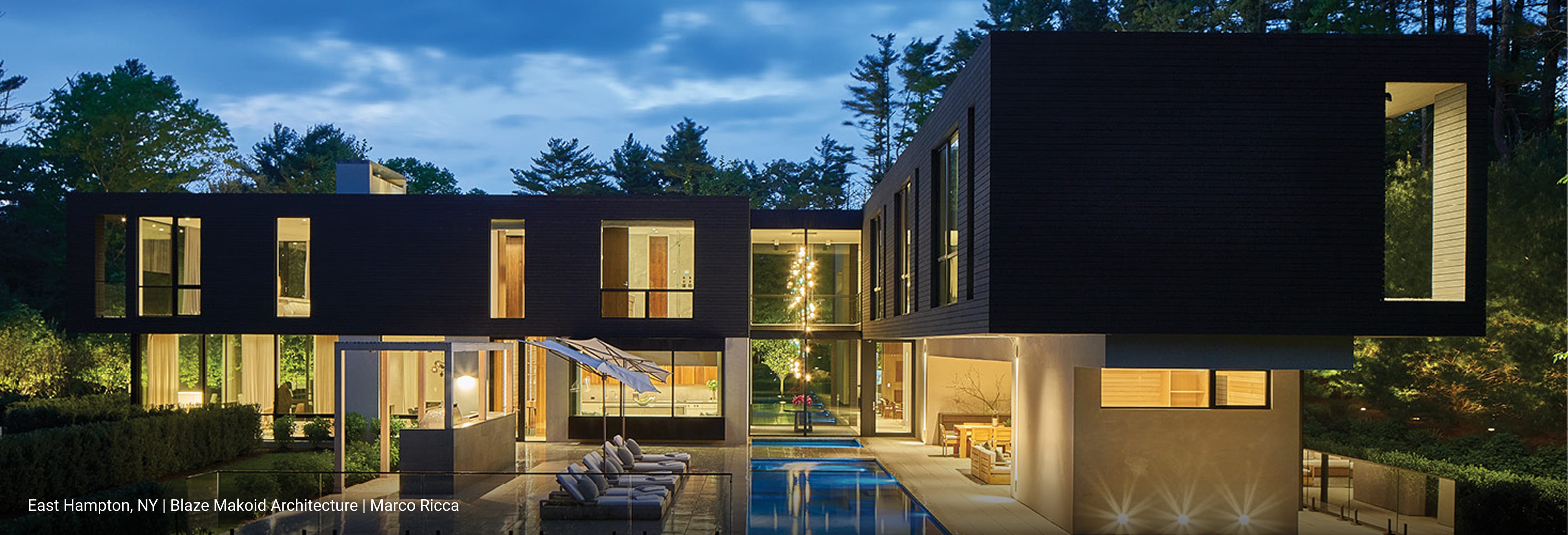 Luxury modern home in featuring dark horizontal wood siding, cantilevered upper level, expansive floor-to-ceiling glass windows, and outdoor pool with lounge chairs illuminated at dusk.