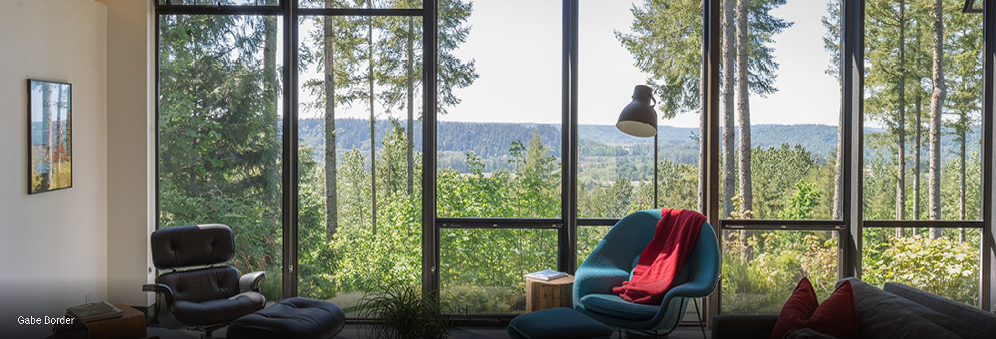 Cozy modern living room with floor-to-ceiling windows offering panoramic forest views, mid-century lounge chairs, vibrant blue armchair with red throw, and natural light-filled interior.