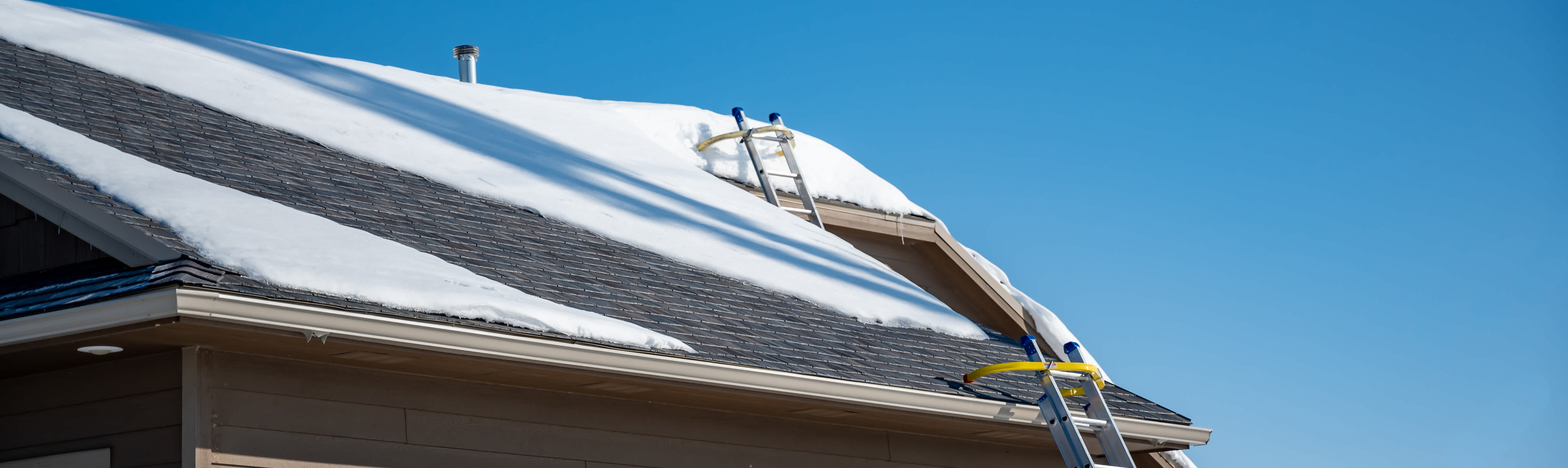 Grey roof with snow blanketed across the surface with a ladder extended up to the rooftop.