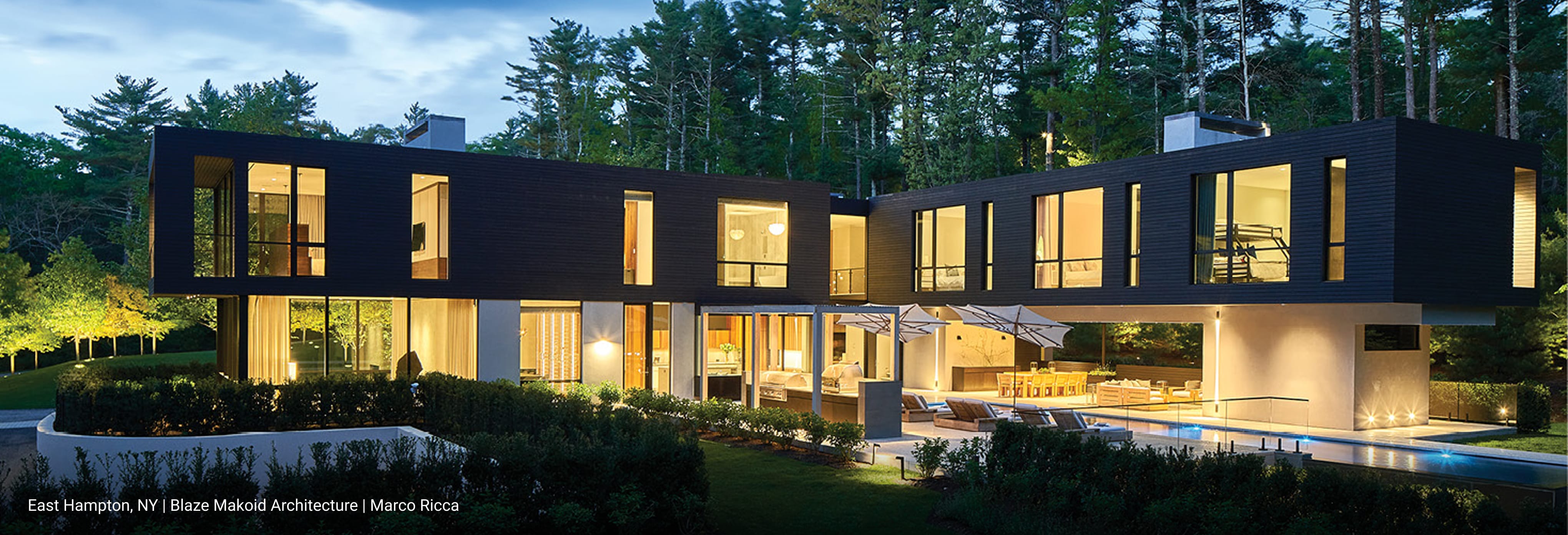 Luxury modern home in East Hampton, New York featuring cantilevered upper level with dark wood siding, expansive floor-to-ceiling glass windows, and open-concept outdoor living space with pool and lounge seating illuminated at dusk.