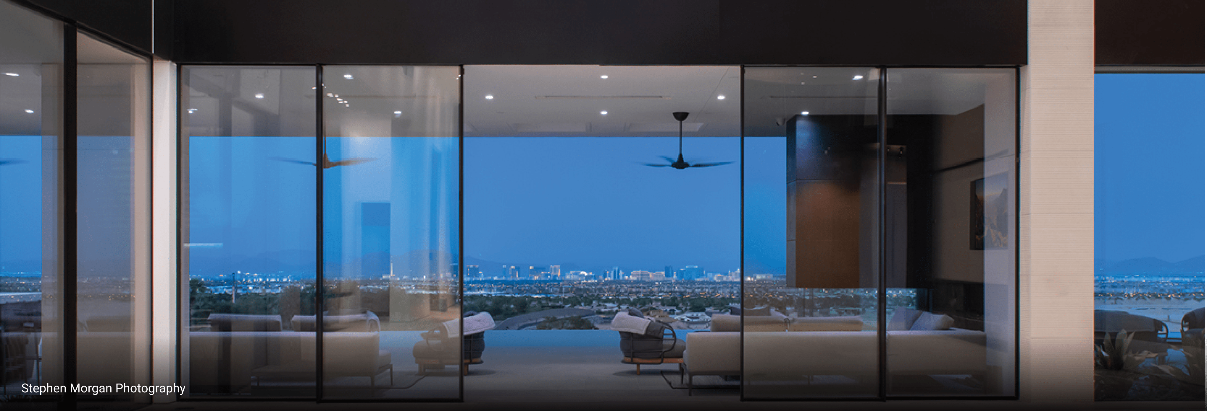 Luxury modern home with floor-to-ceiling glass walls offering panoramic city views at dusk, featuring sleek minimalist interior and outdoor seating area.