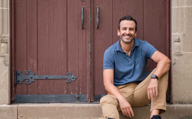 Chad Esslinger wearing a blue henley shirt and khaki pants sitting in front of a reddish brown antique exterior door.