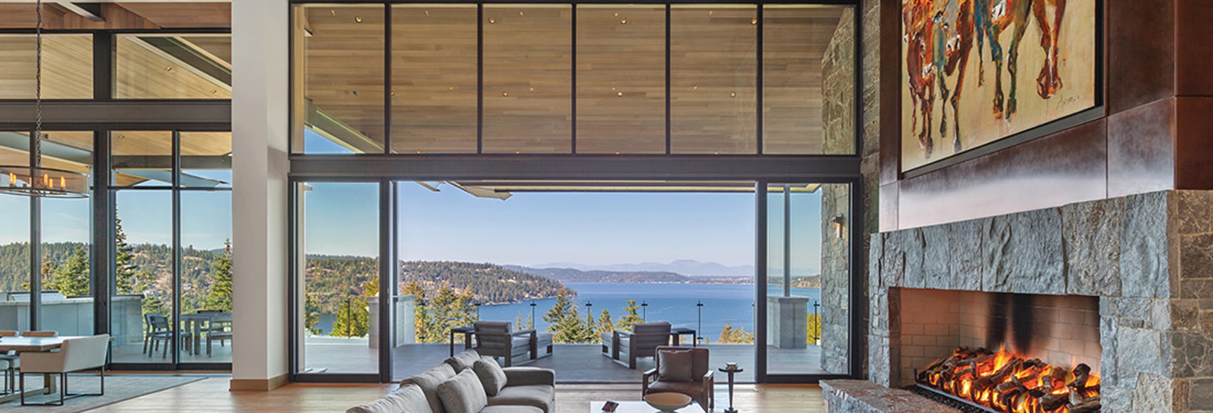 Luxury open-concept living room with stone fireplace, large sectional sofa, and floor-to-ceiling glass doors opening to a terrace with panoramic lake and mountain views.