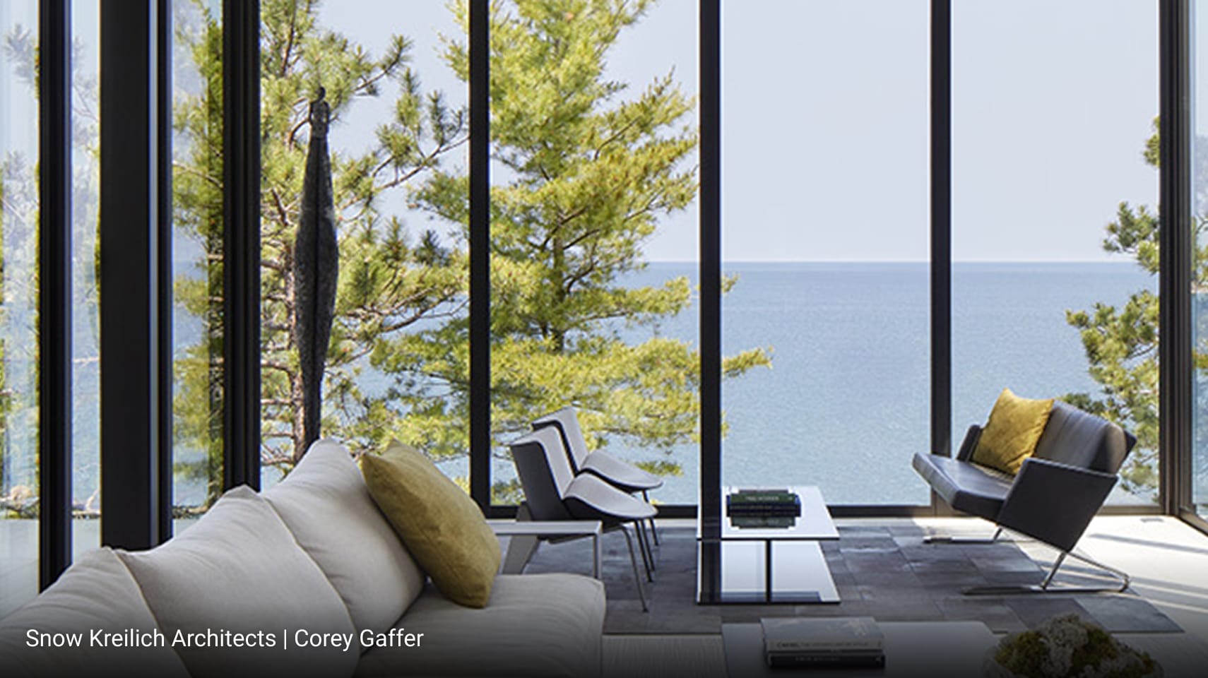 Modern living room with floor-to-ceiling glass walls offering panoramic lake views, minimalist furniture including light beige sofa and black lounge chair, sleek coffee tables, and natural stone accent wall.