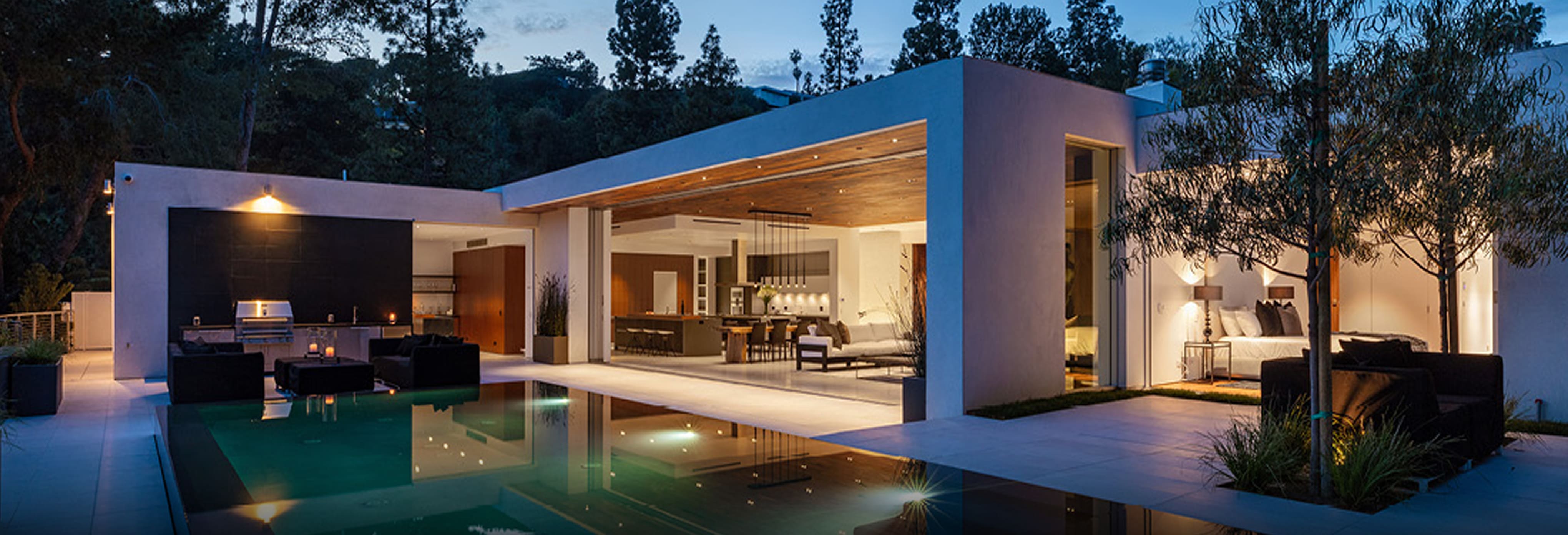 Luxury modern home with open-concept design featuring floor-to-ceiling sliding glass doors, white stucco exterior, wood-paneled ceiling, and outdoor living space with illuminated swimming pool and lounge seating at dusk.