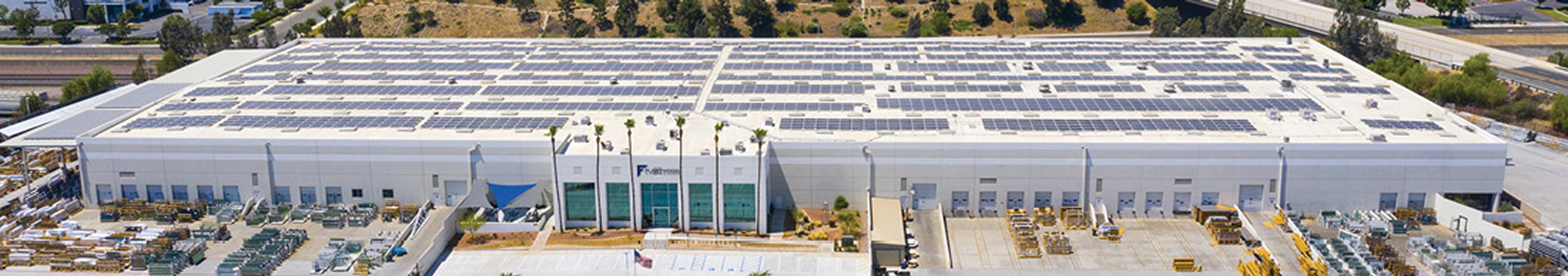 Fleetwood manufacturing facility with expansive rooftop solar panel installation, large white industrial building, and organized outdoor storage area for architectural products and materials.