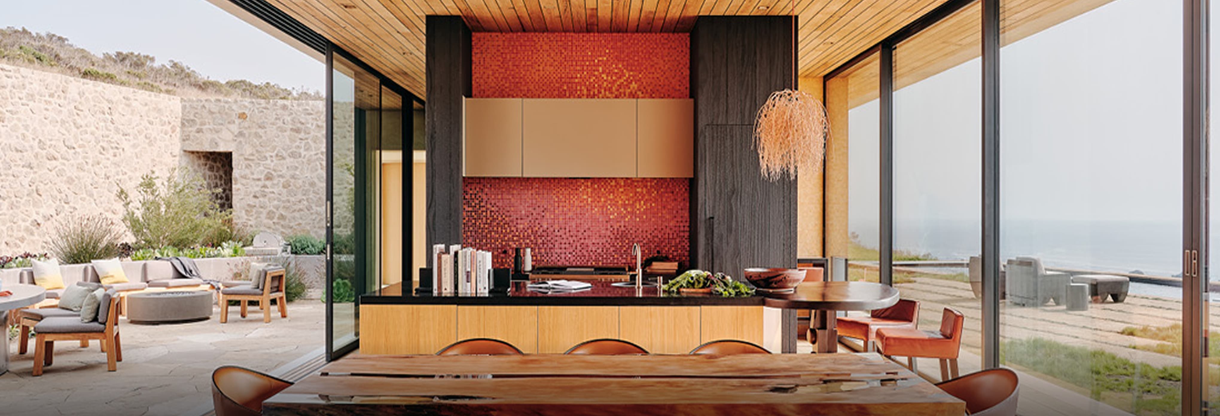 Luxury open-concept kitchen and dining area with natural wood ceiling, large glass sliding doors opening to ocean-view patio, red mosaic tile backsplash, black countertop island, and rustic wooden dining table with leather chairs.