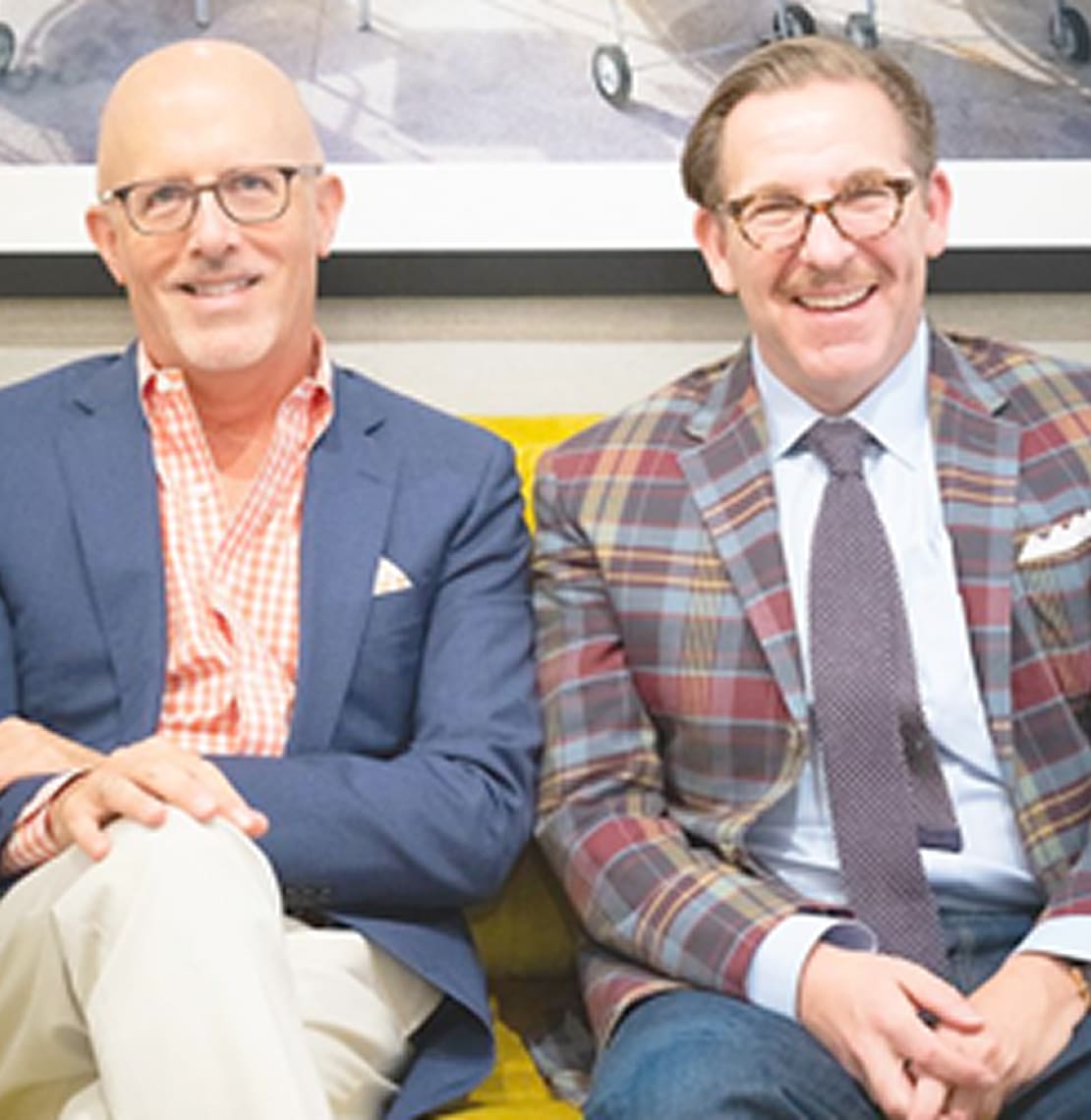 Interior setting featuring two individuals seated on a mustard-yellow sofa, dressed in tailored blazers with patterned shirts and ties, against a backdrop of framed artwork depicting an airplane.