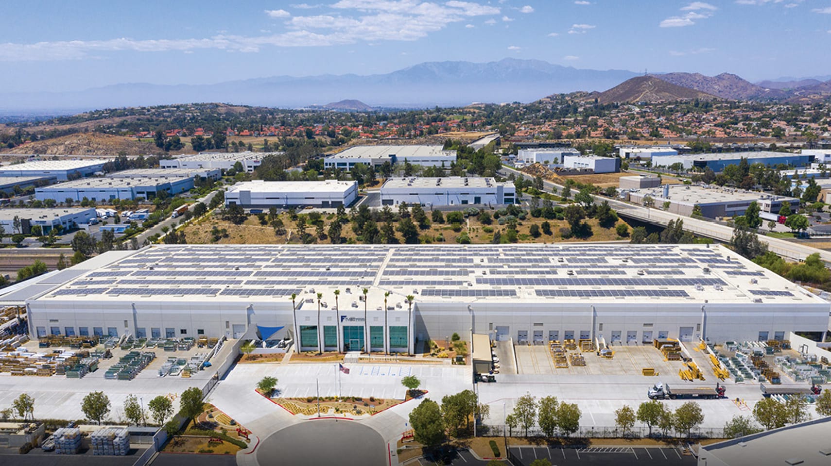 Aerial view of Fleetwood manufacturing facility with expansive rooftop solar panel installation, large white industrial building, organized outdoor storage areas, and surrounding business park landscape under clear skies.