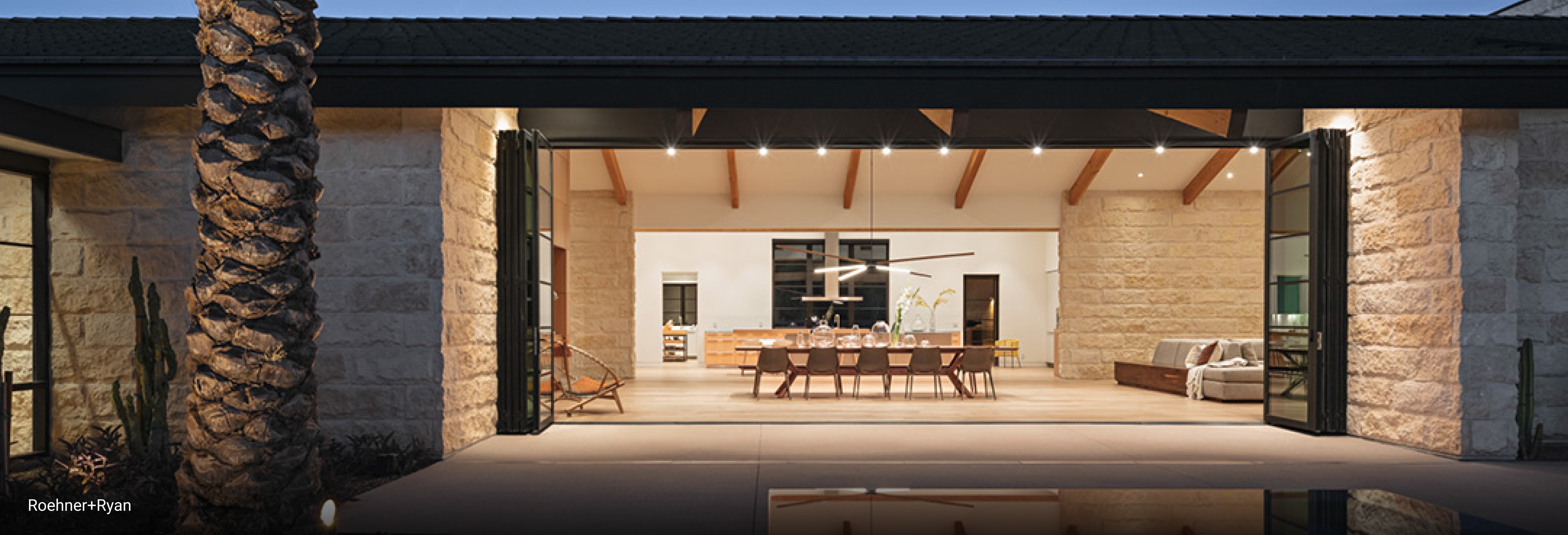 Luxury open-concept dining and living area with exposed wood ceiling beams, stone walls, large folding glass doors opening to outdoor patio, and modern chandelier above a long wooden dining table.