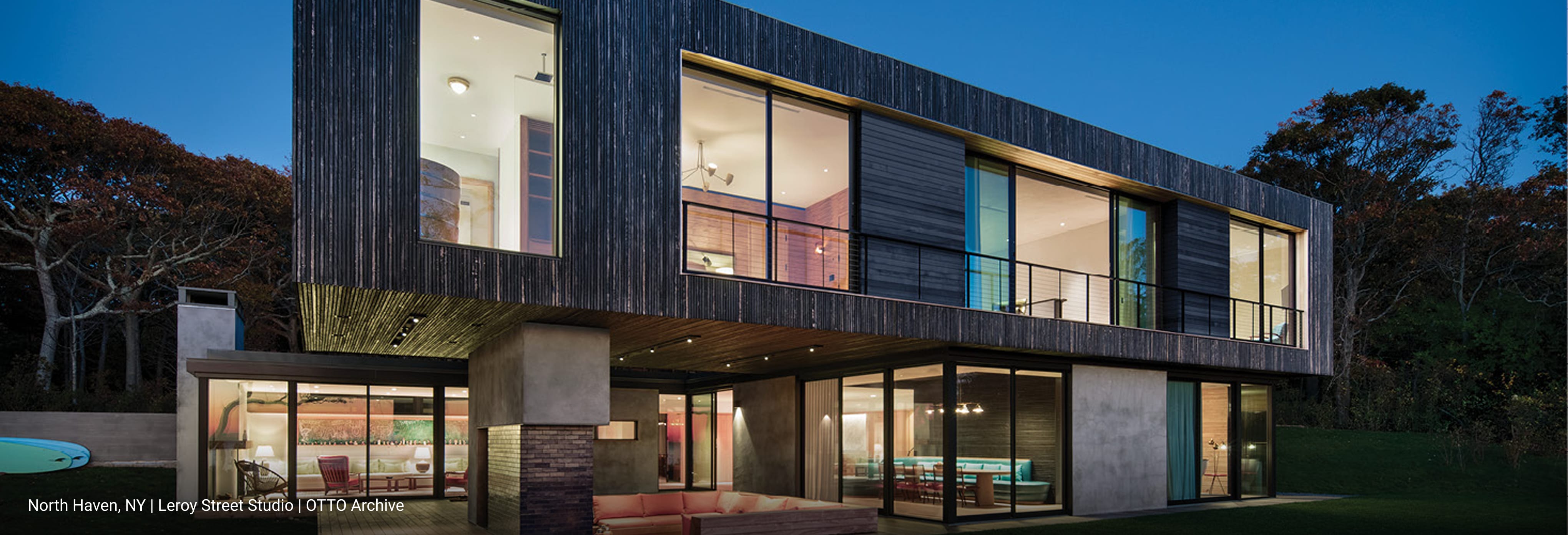 Contemporary two-story home in featuring dark vertical wood siding, cantilevered upper level with floor-to-ceiling glass windows, and open-concept lower level with sliding glass doors overlooking landscaped lawn.