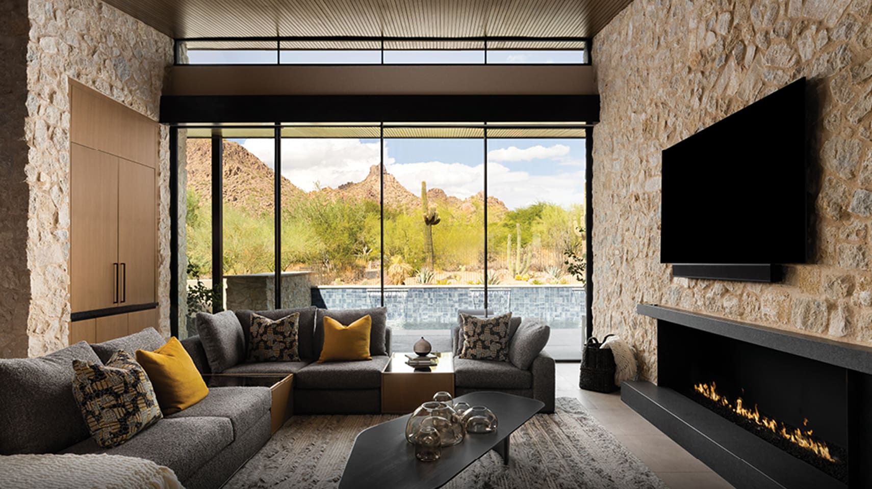 Luxury living room with natural stone walls and floor-to-ceiling glass windows offering panoramic desert mountain views, modern gray sectional sofa with patterned and yellow accent pillows, sleek black fireplace, and minimalist decor.